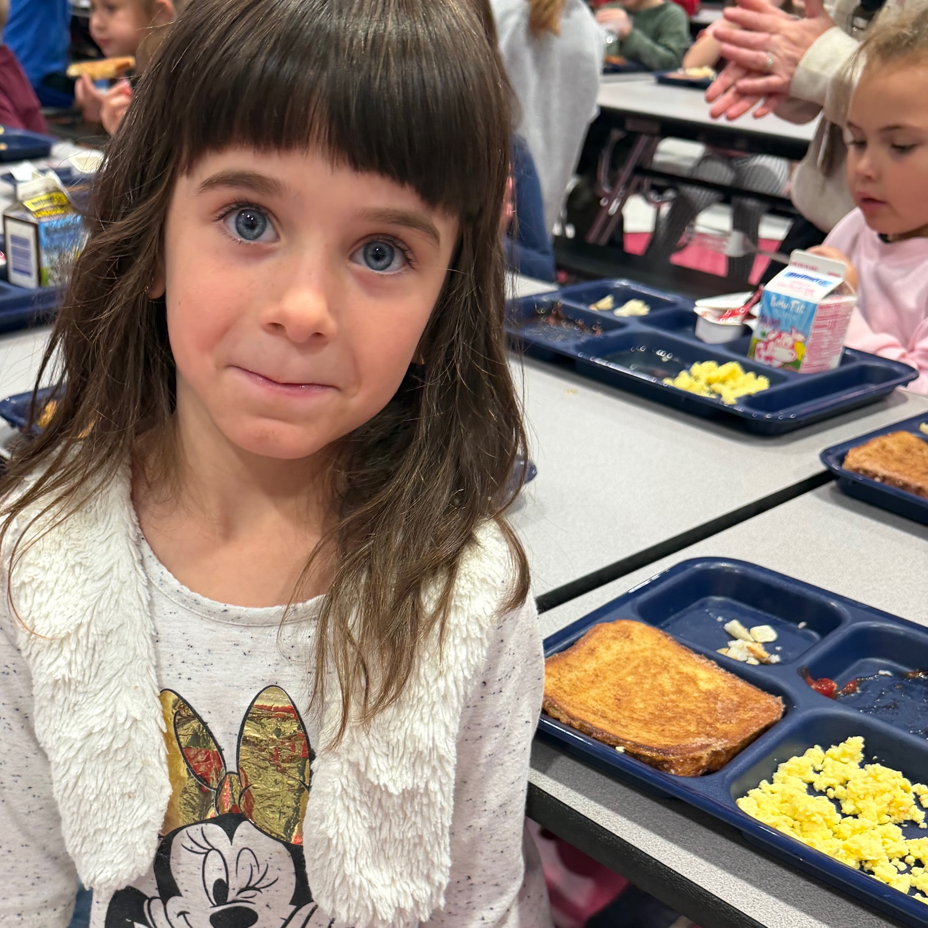 Two children sit at a cafeteria table with lunch trays and other children sitting at tables in the background.
