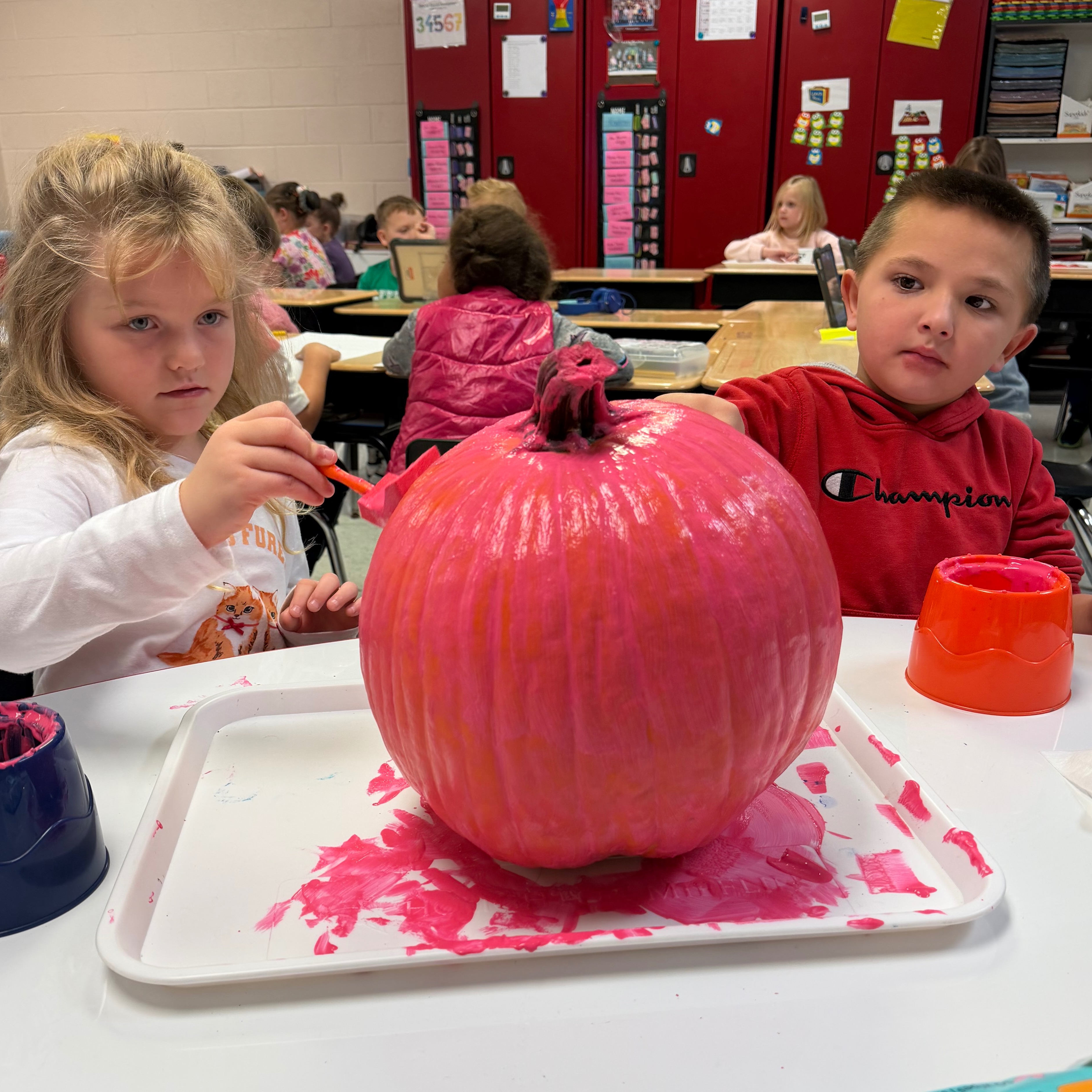 Two kids painting a large pumpkin on a table. Paint tray has red paint. Background shows other kids and tables.