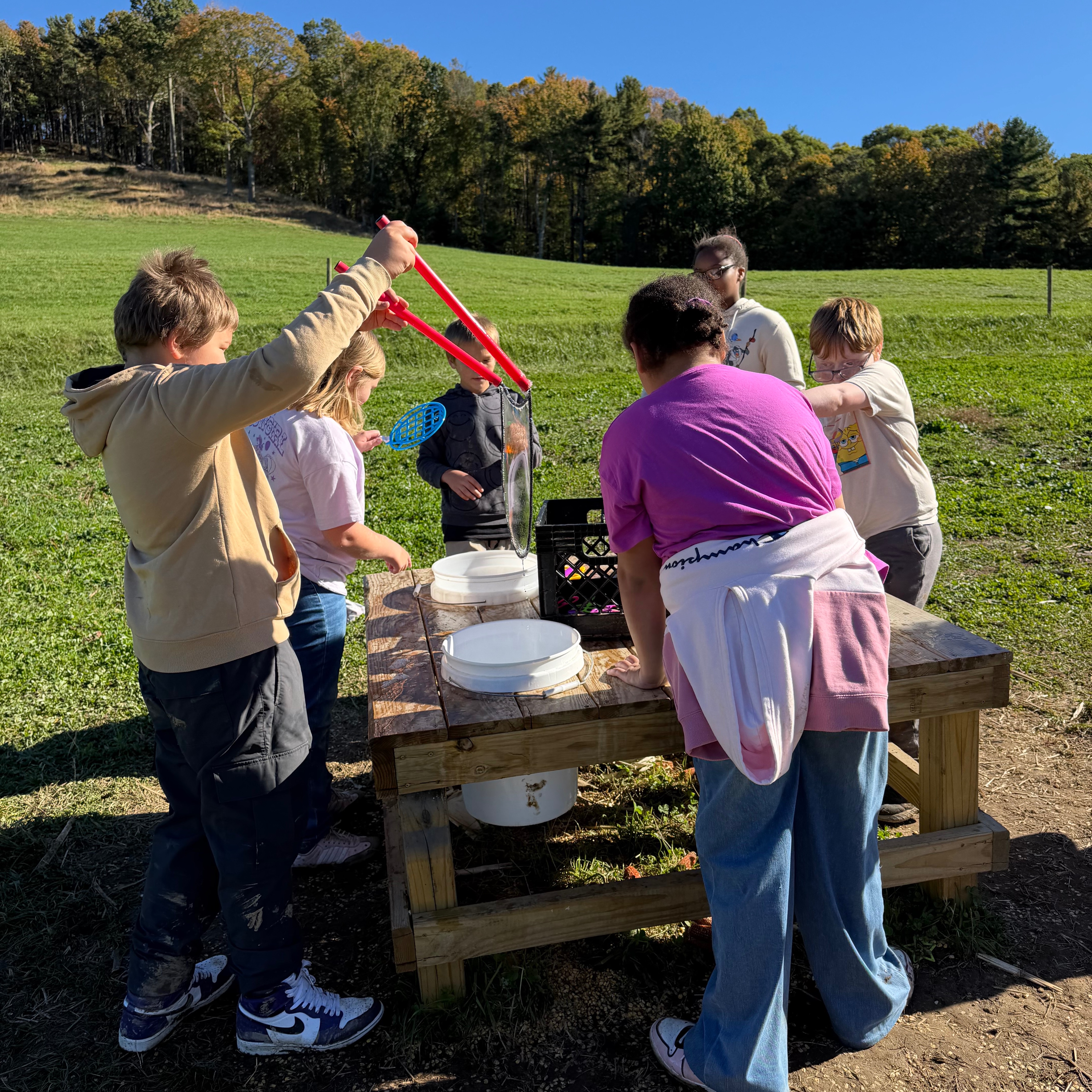 People around a table outdoors, some using tools and others standing with bowls. Green field and trees in the background.