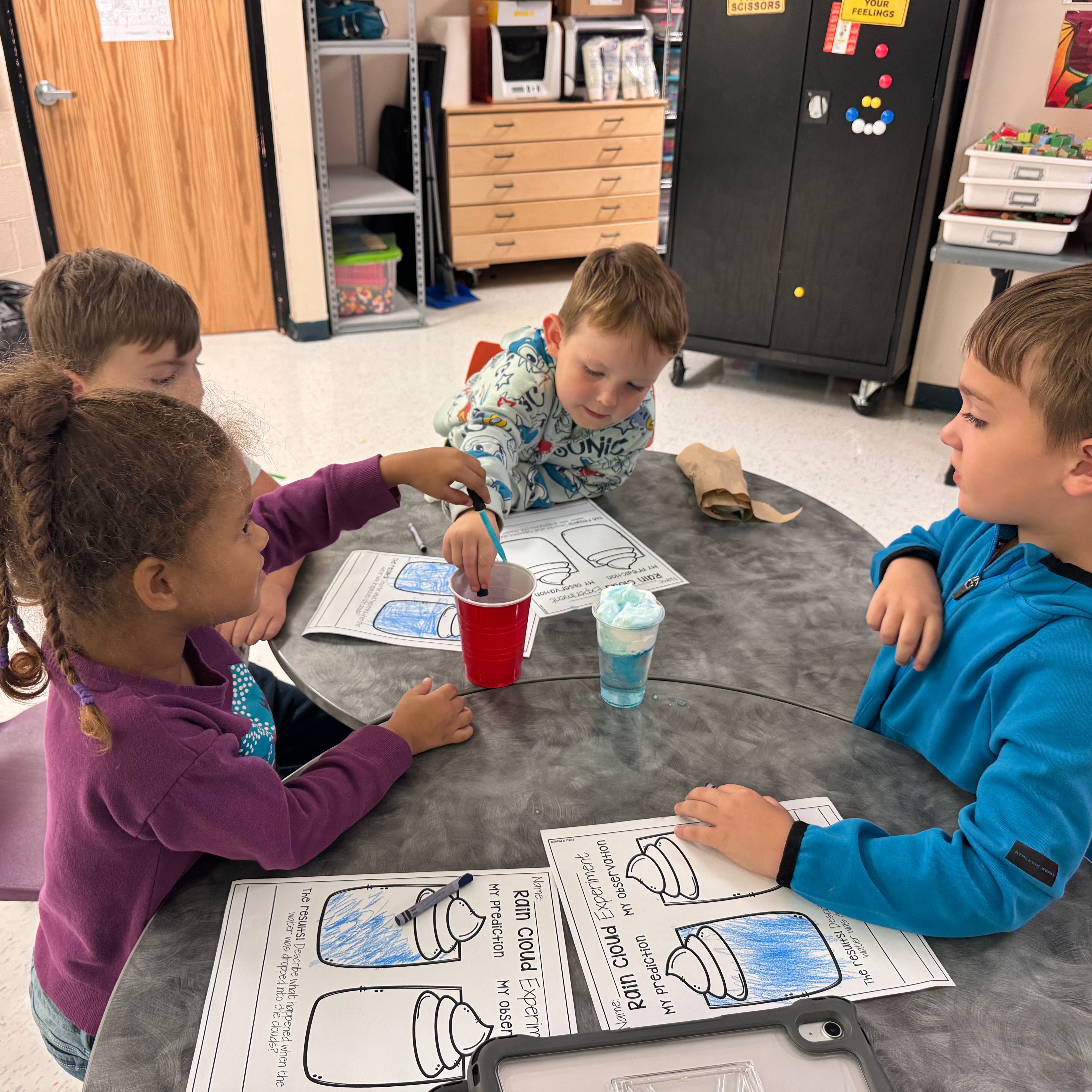 Four children seated around a table; one girl holds a cup, another girl touches it; two boys listen