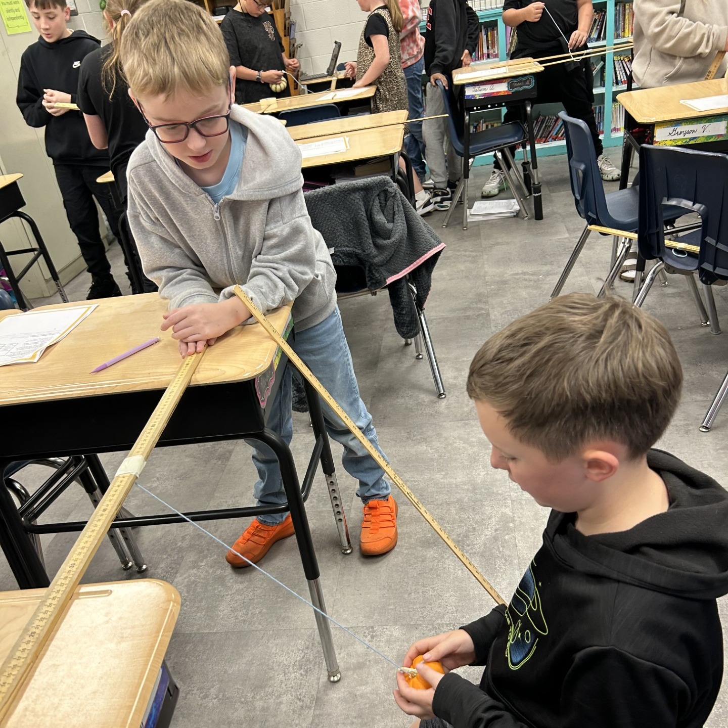 Two boys work together to straighten a long rope, one sitting, one standing, with desks and chairs in a classroom.