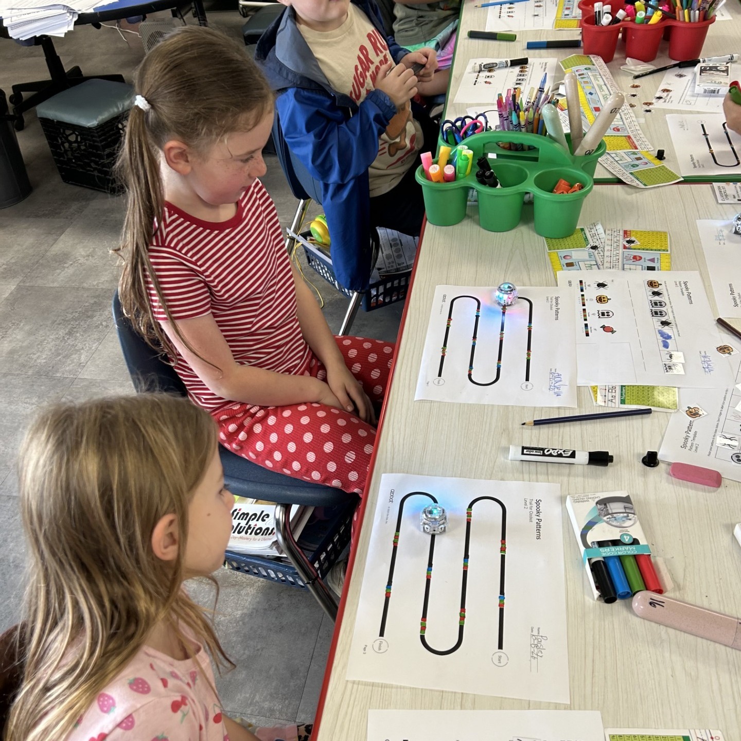 Students sit at tables, one wearing a red and white polka-dot shirt. The table has papers with drawings, colored pencils, and markers.