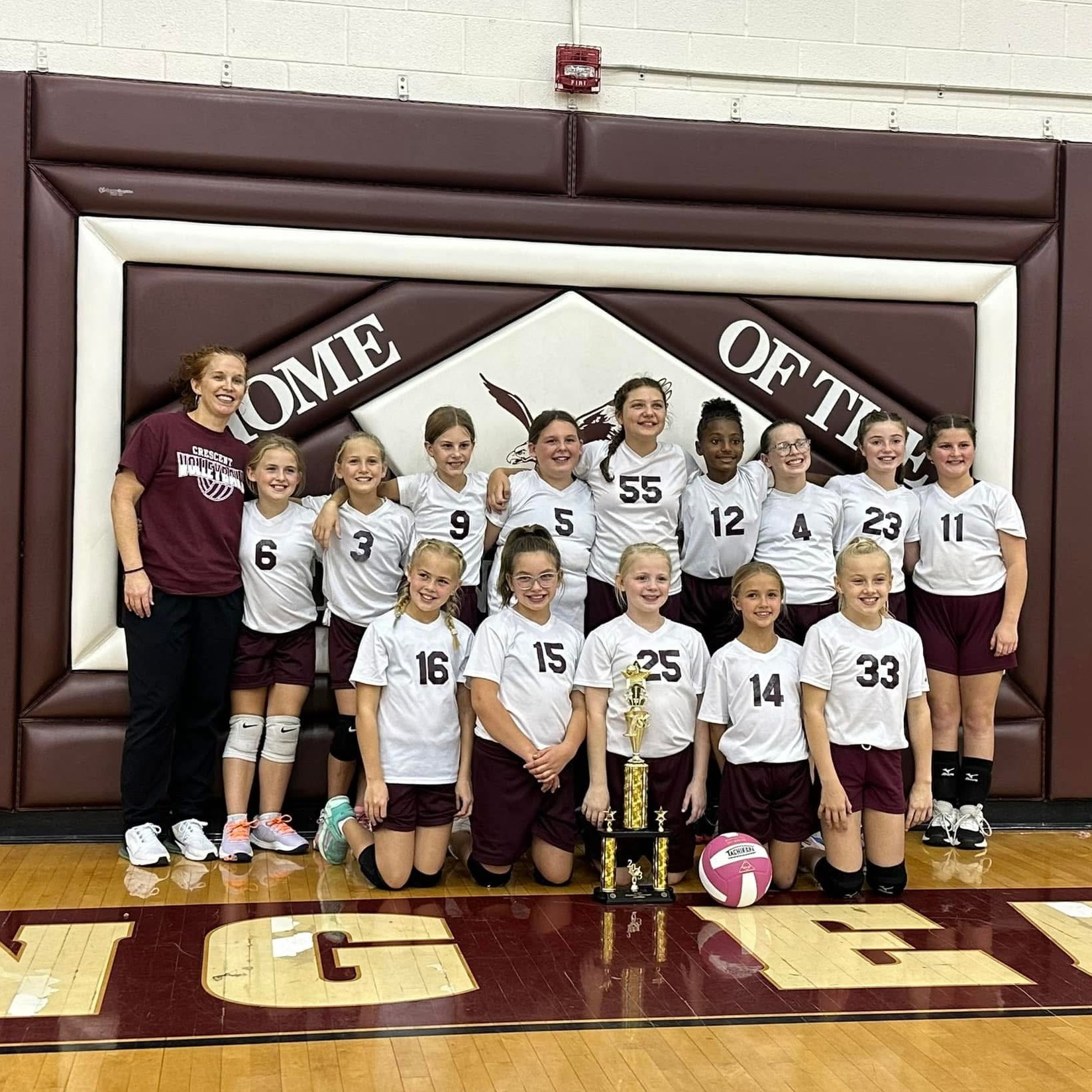 Young girls in white and maroon uniforms posing with trophies and a volleyball on a basketball court.