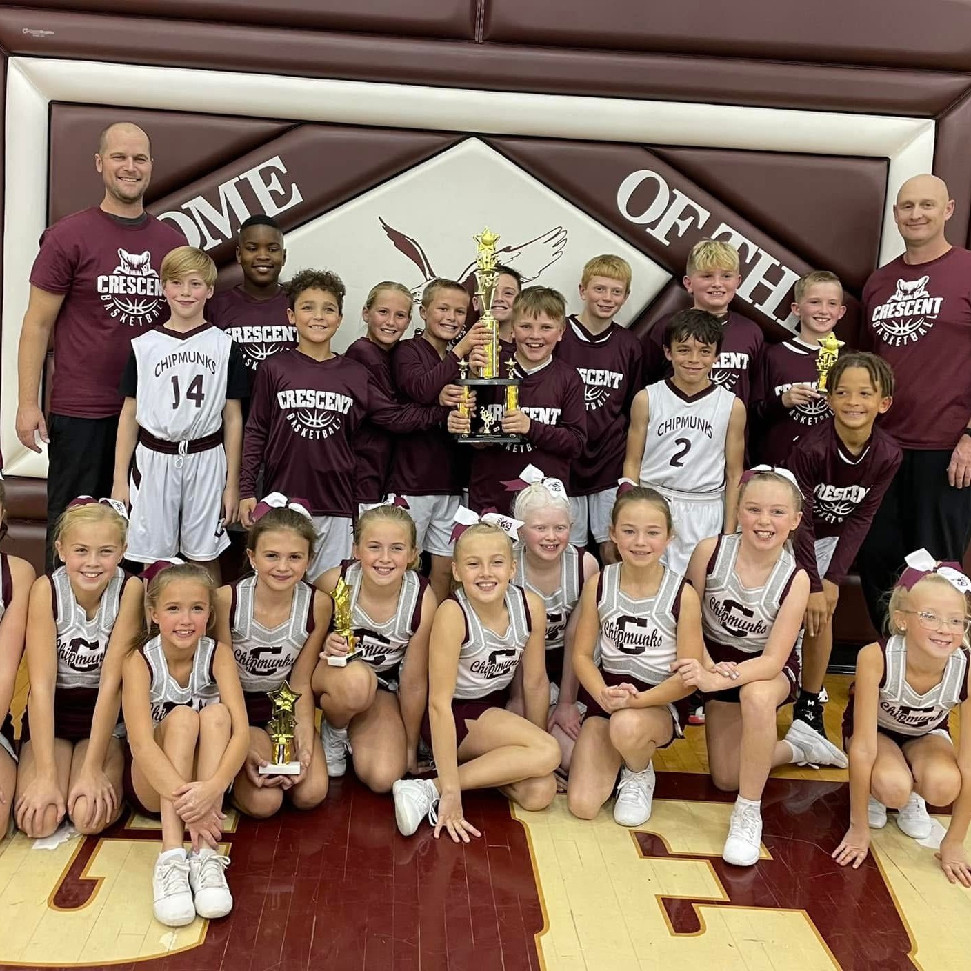 Basketball team poses for photo holding trophies, with school motto on a wall in the background.