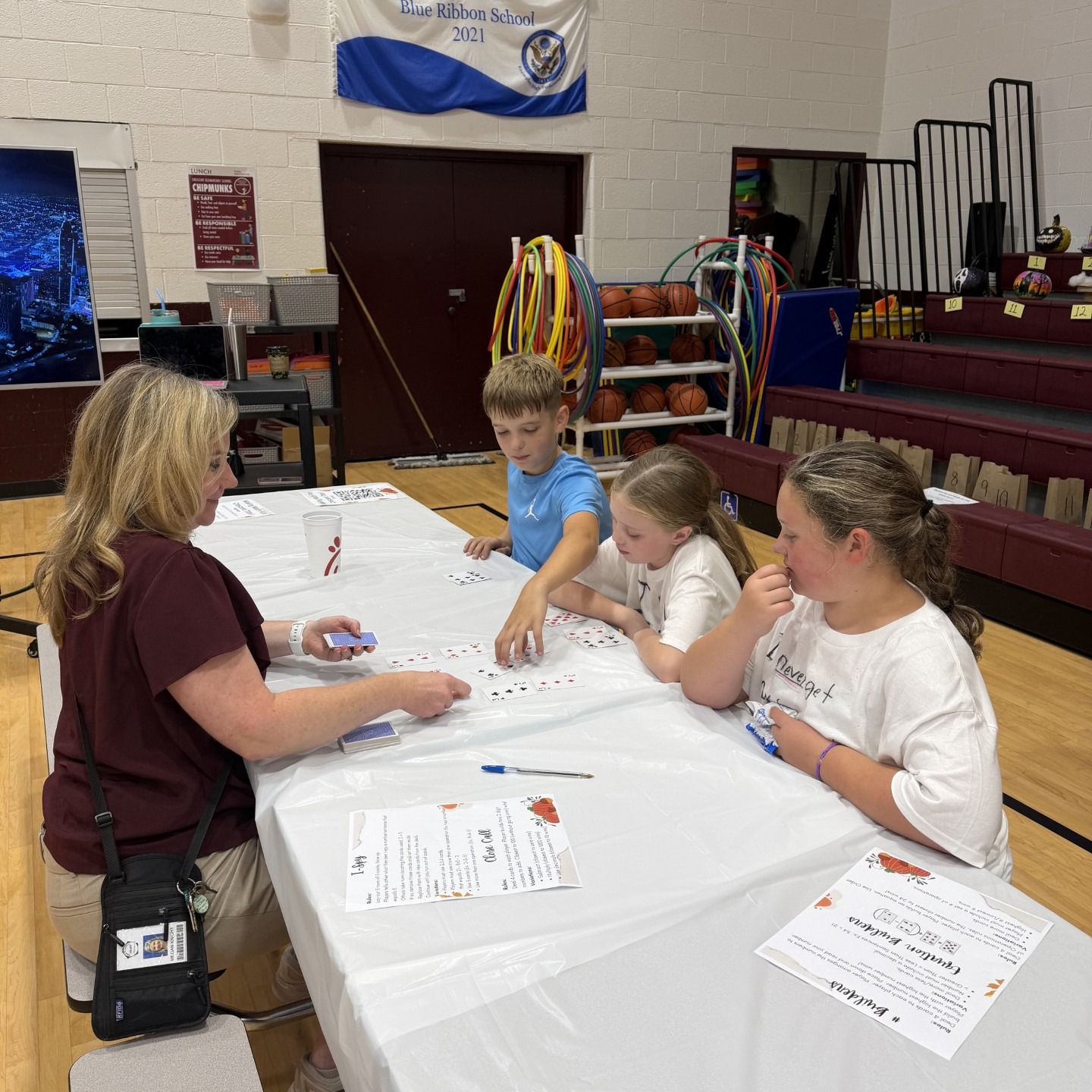 Three students and an adult seated at a long table with papers, pencils, and a bag.