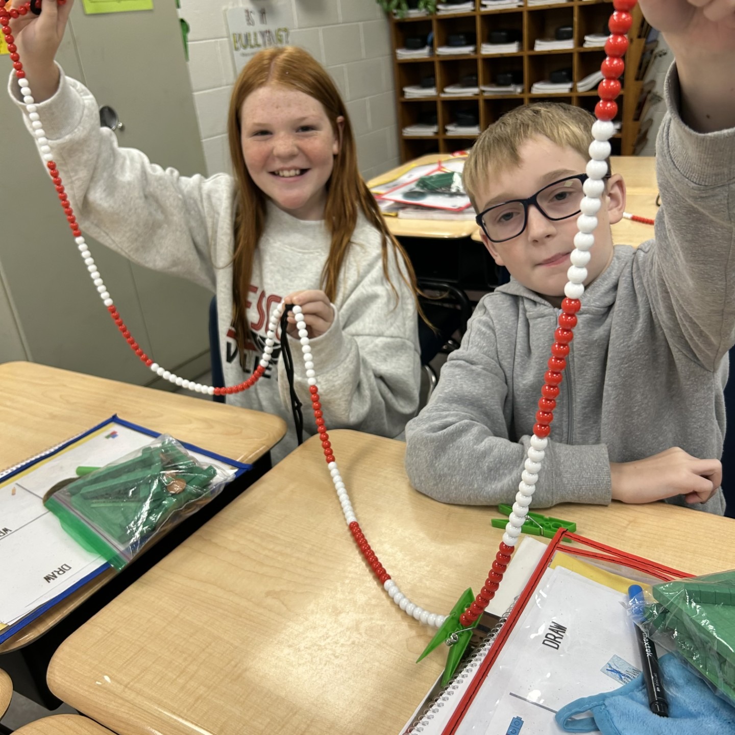 Two students sit at a desk holding up red and white beaded strings. Papers, pencils, and a plant are on the desk.