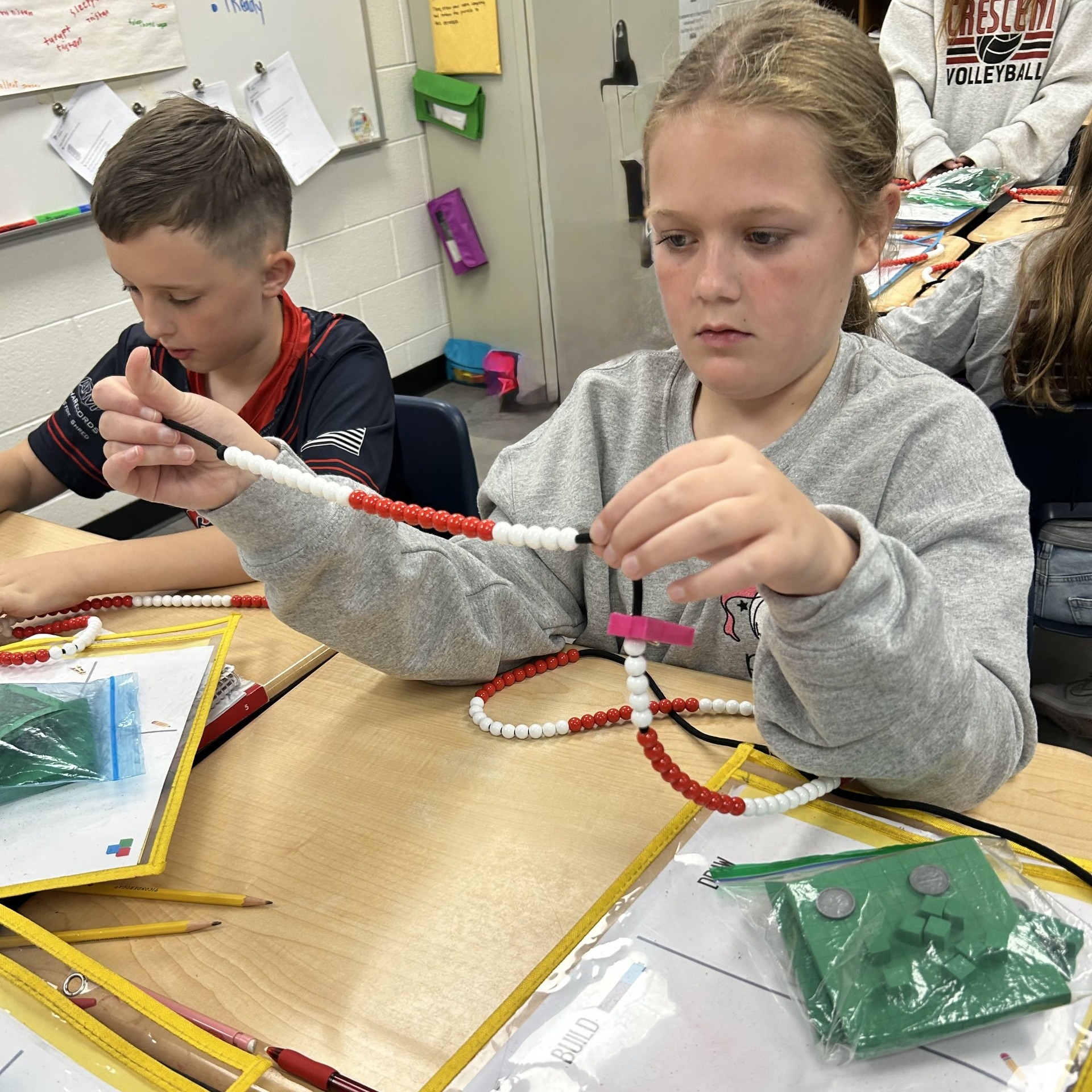 Two students work on a craft project using colored beads at a school table.