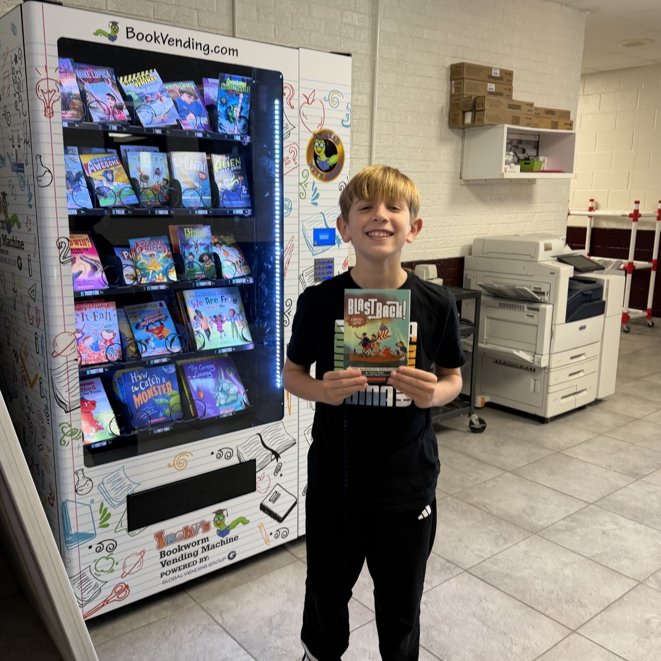Boy in black shirt holding a book in front of a white book vending machine.