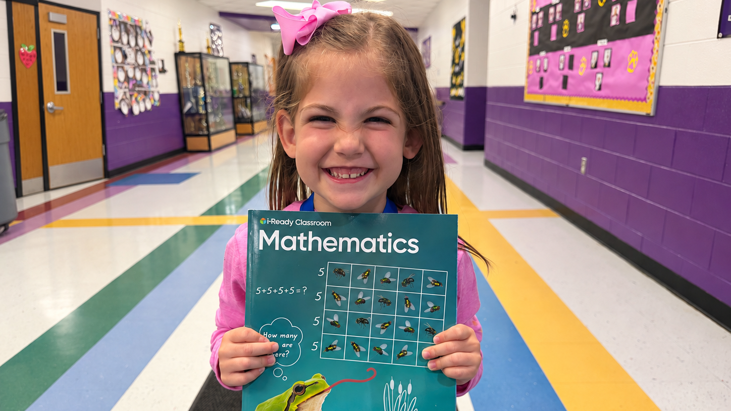 Girl holding a math book, smiling, with a frog on the cover, in a school hallway.
