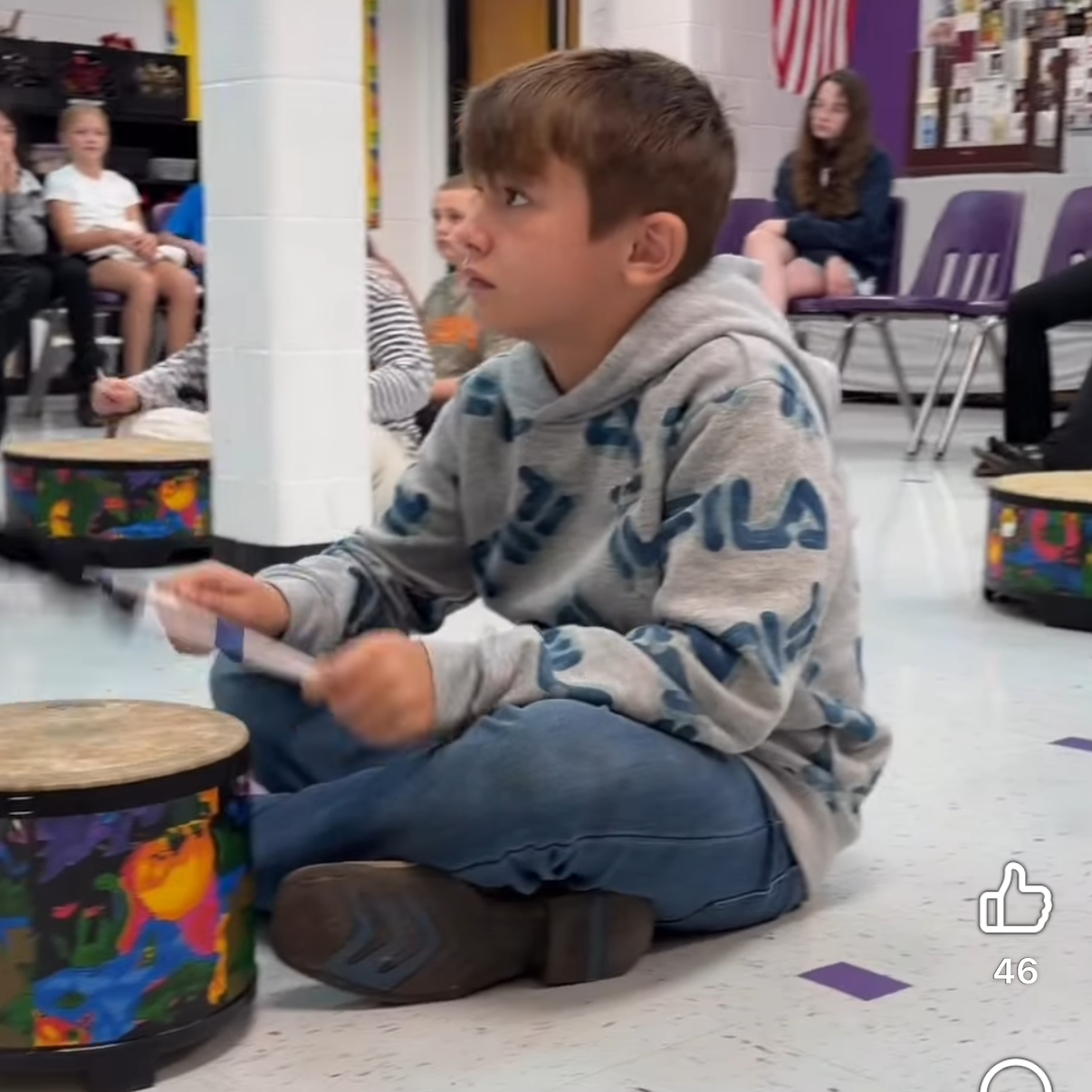 Boy sitting on floor with drums in front of him, looking at another boy, with other children seated nearby.