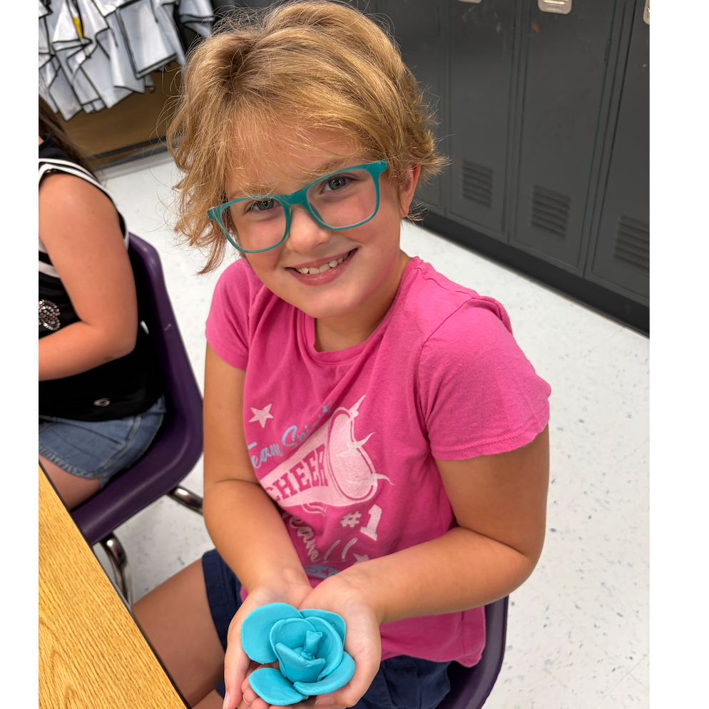 Blonde girl in glasses holds blue rose-shaped object. Lockers and another student visible in background.