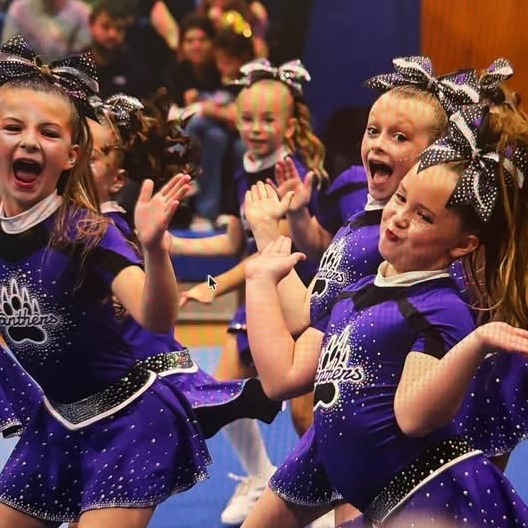 Cheerleaders wearing purple uniforms with white trim perform on a gym floor, some clapping hands.