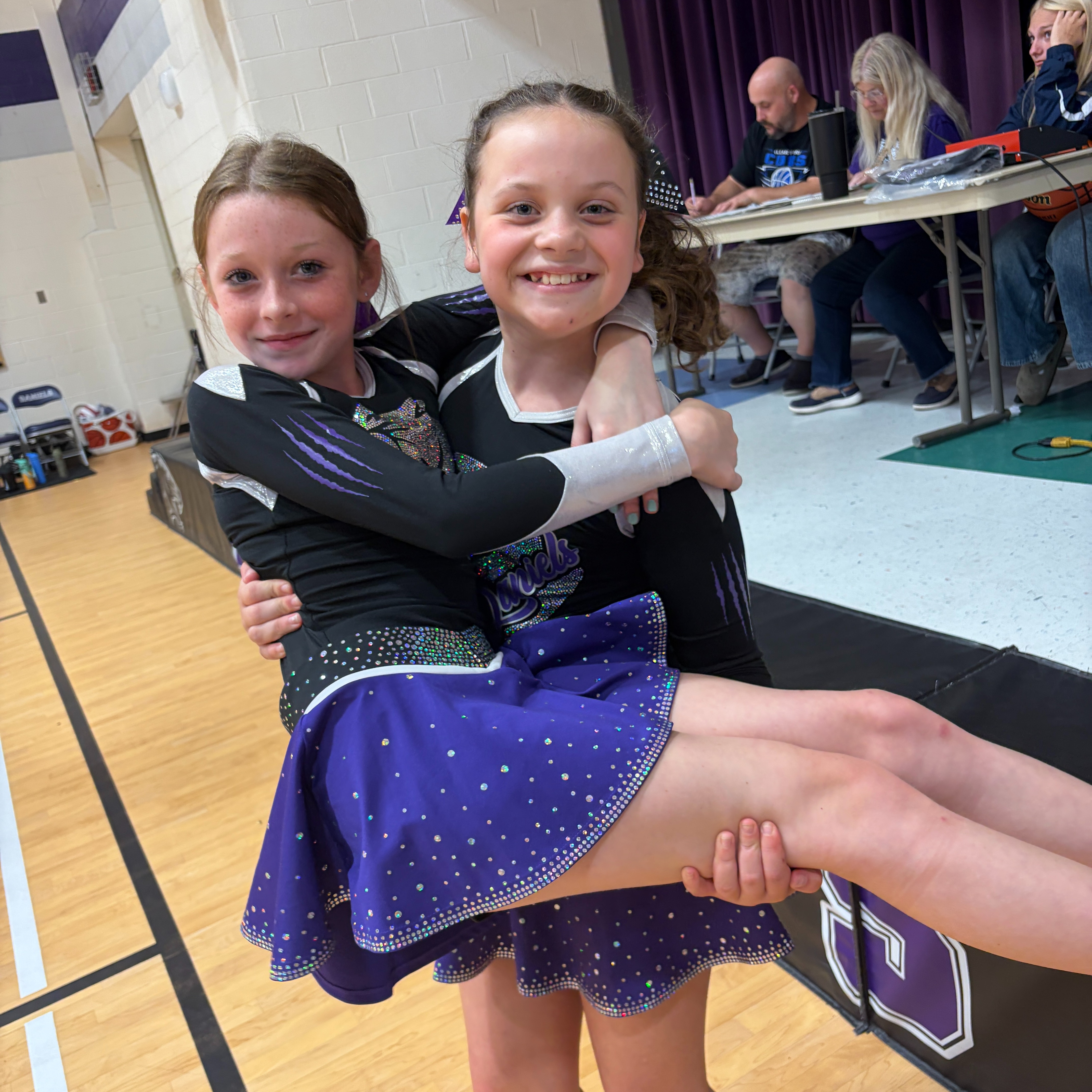 Two girls in matching uniforms hug, one lifted by the other in a gym. Behind them, people sit at a table.