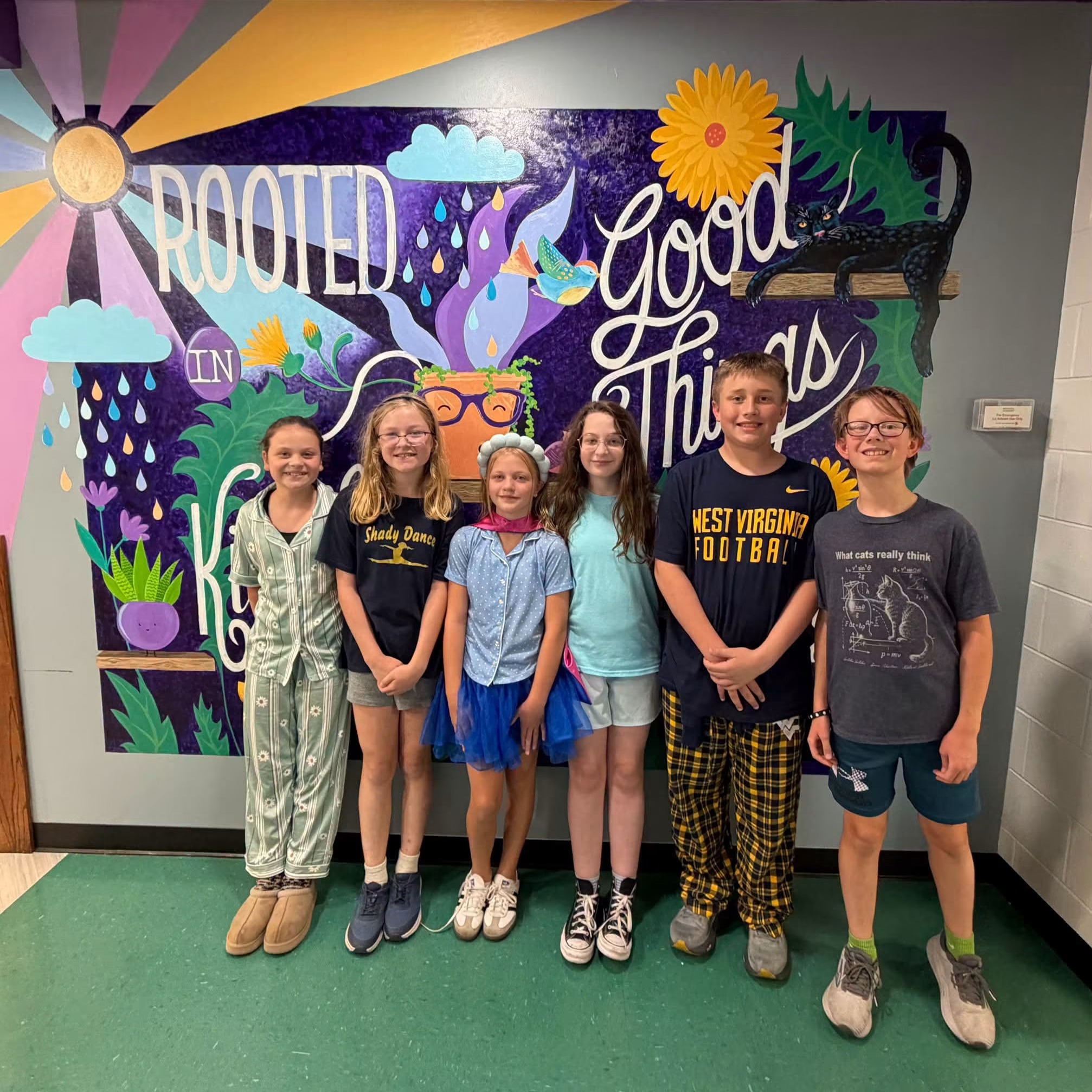 Seven students standing in front of a mural with plants, flowers, and sun. They wear casual clothes.