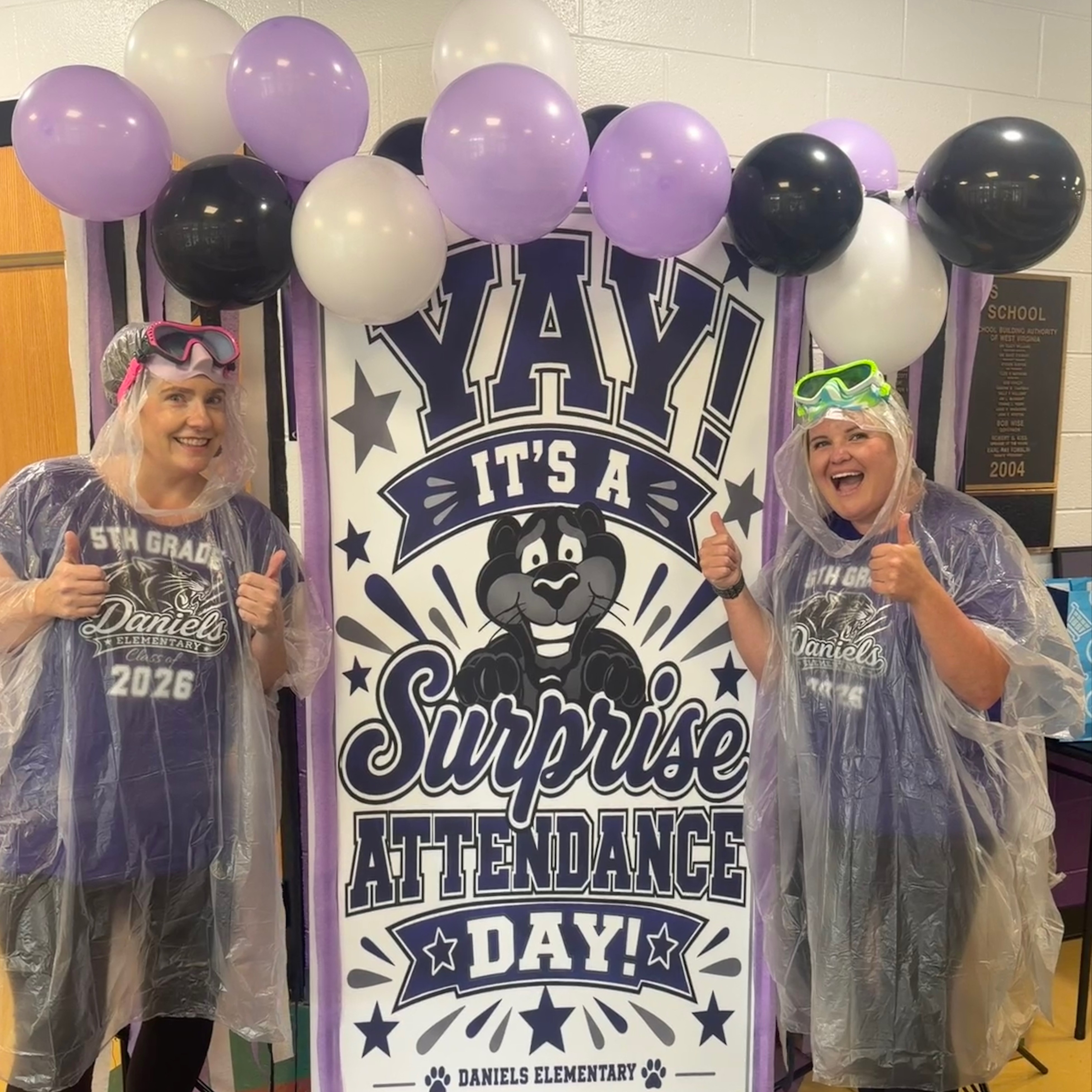 Two women in rain ponchos stand by a banner with balloons and text that reads "YAY! It's a Surprise Attendance Day!"