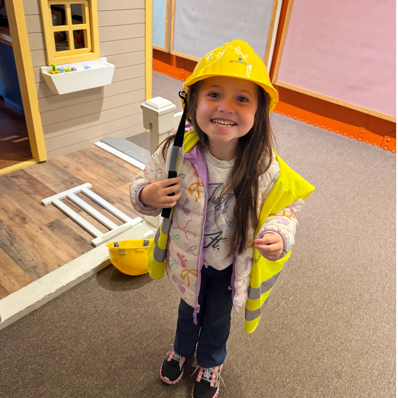 A girl in a hard hat and vest, smiling, standing in a play area with a toy ladder and a playhouse.