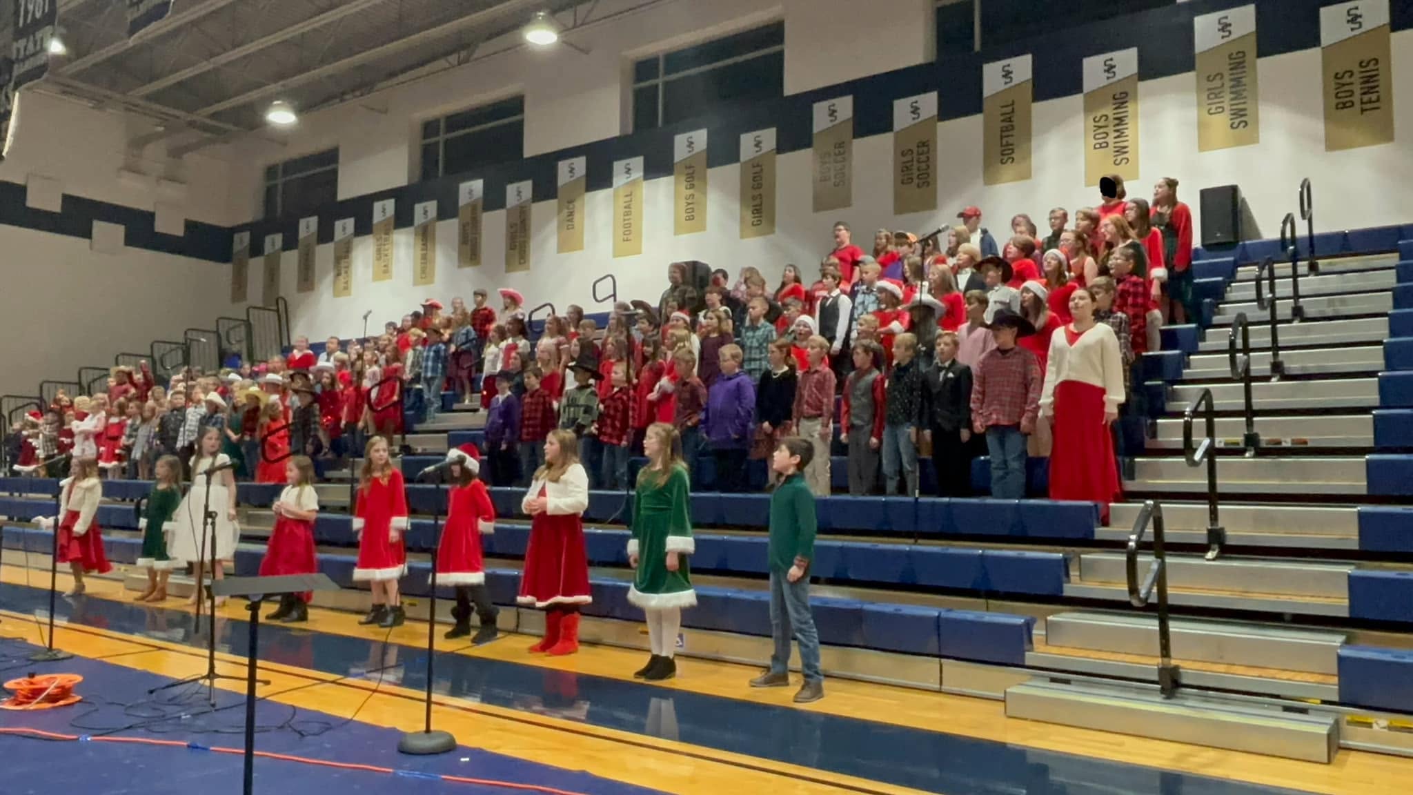 A group of people dressed in holiday attire on bleachers in a gymnasium, with some on stage.