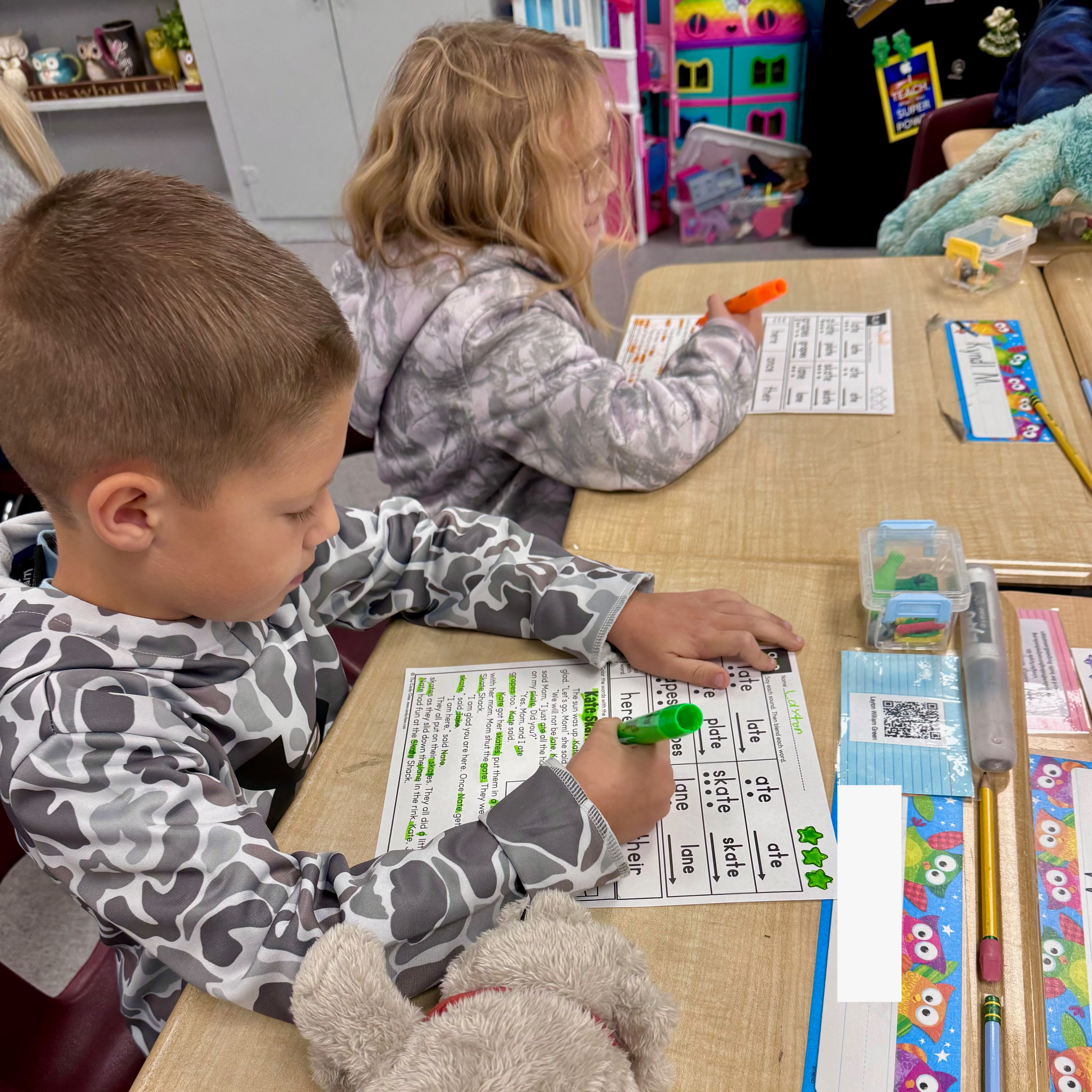 Two children sit at a desk, working on a worksheet with green markers. A stuffed animal sits nearby.