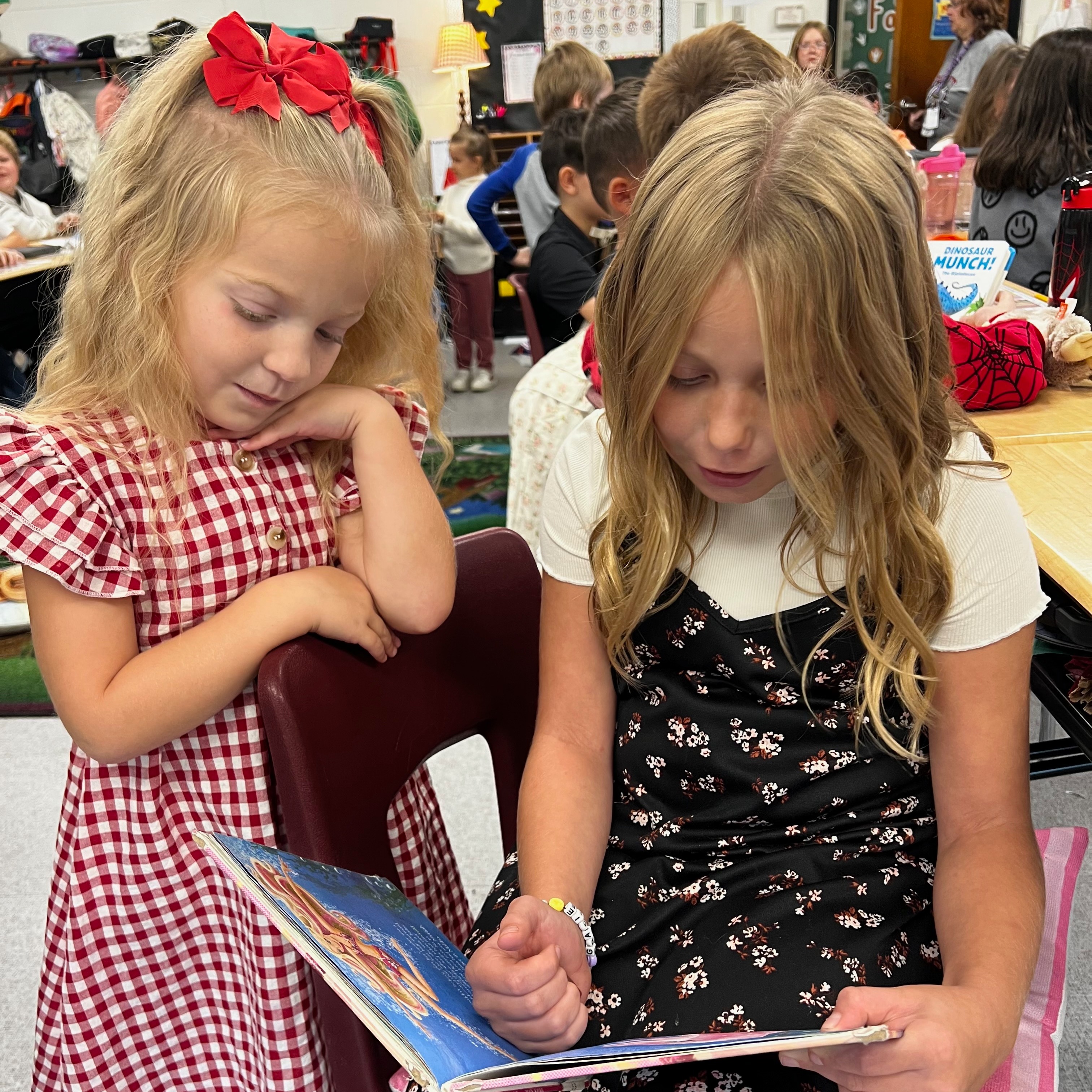Two girls in a classroom, one reading a book to another sitting in a chair.