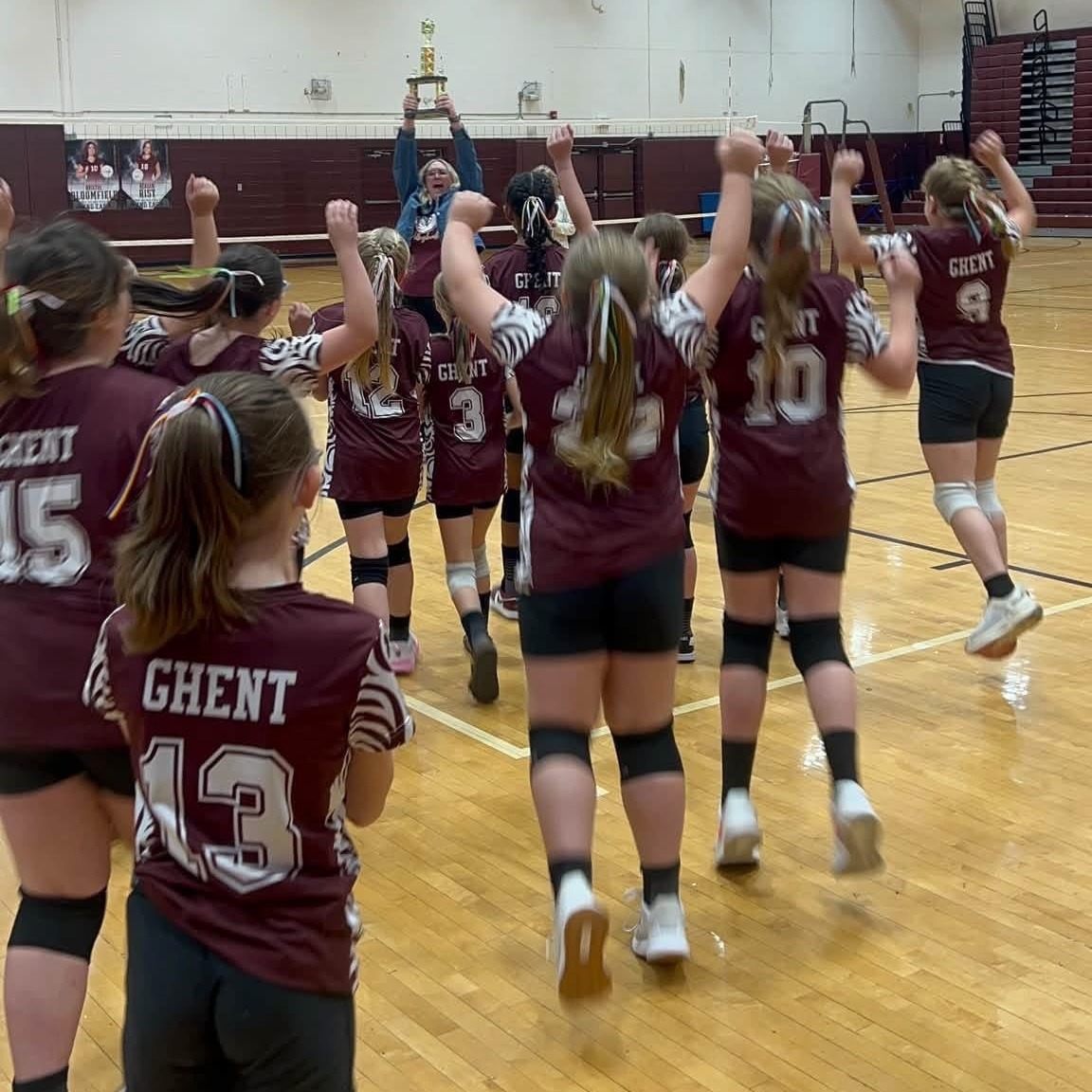 Volleyball team celebrates victory, holding a trophy, on a basketball court with hardwood floor.