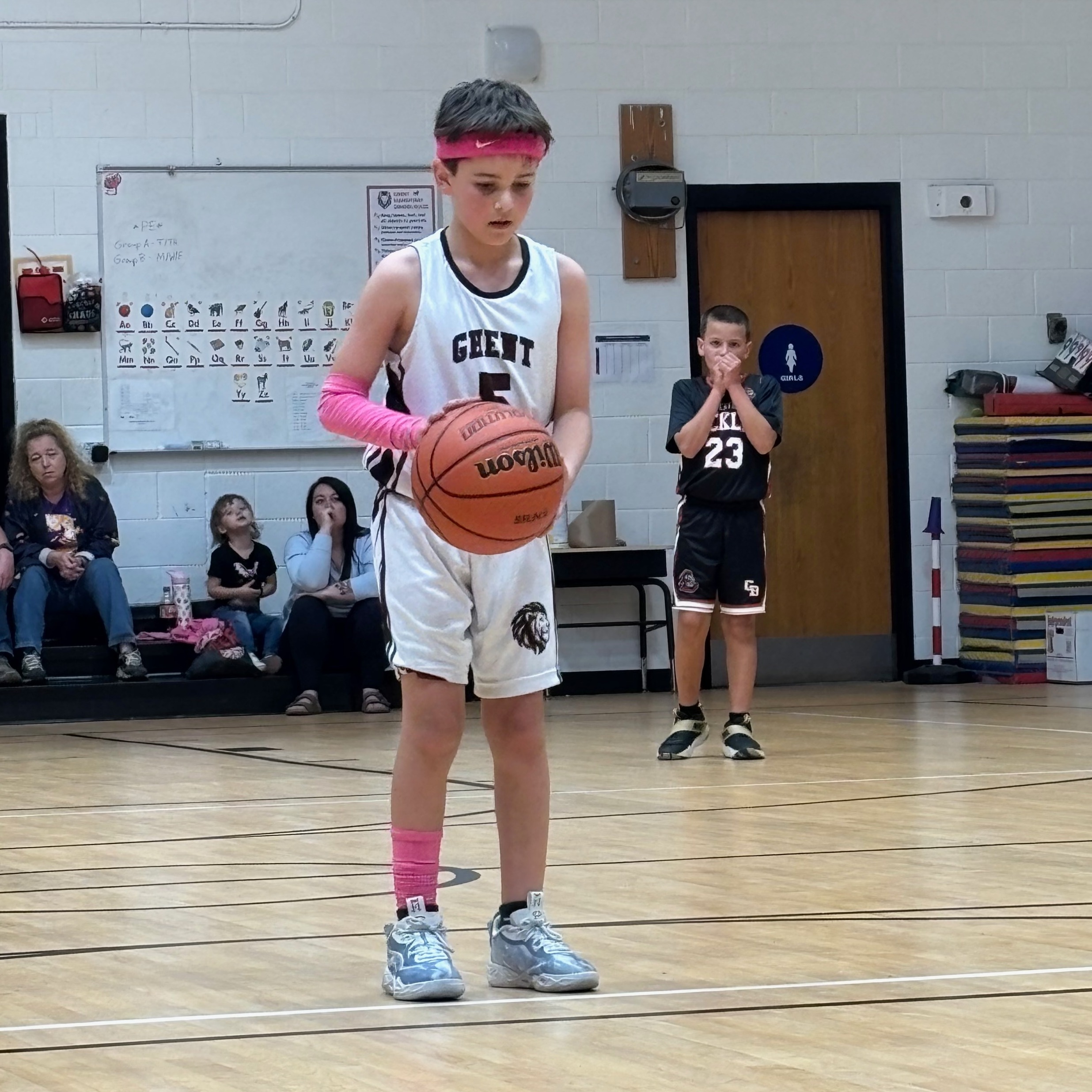 A boy holds a basketball while standing on a basketball court. A player stands behind him. Spectators sit on a bench.
