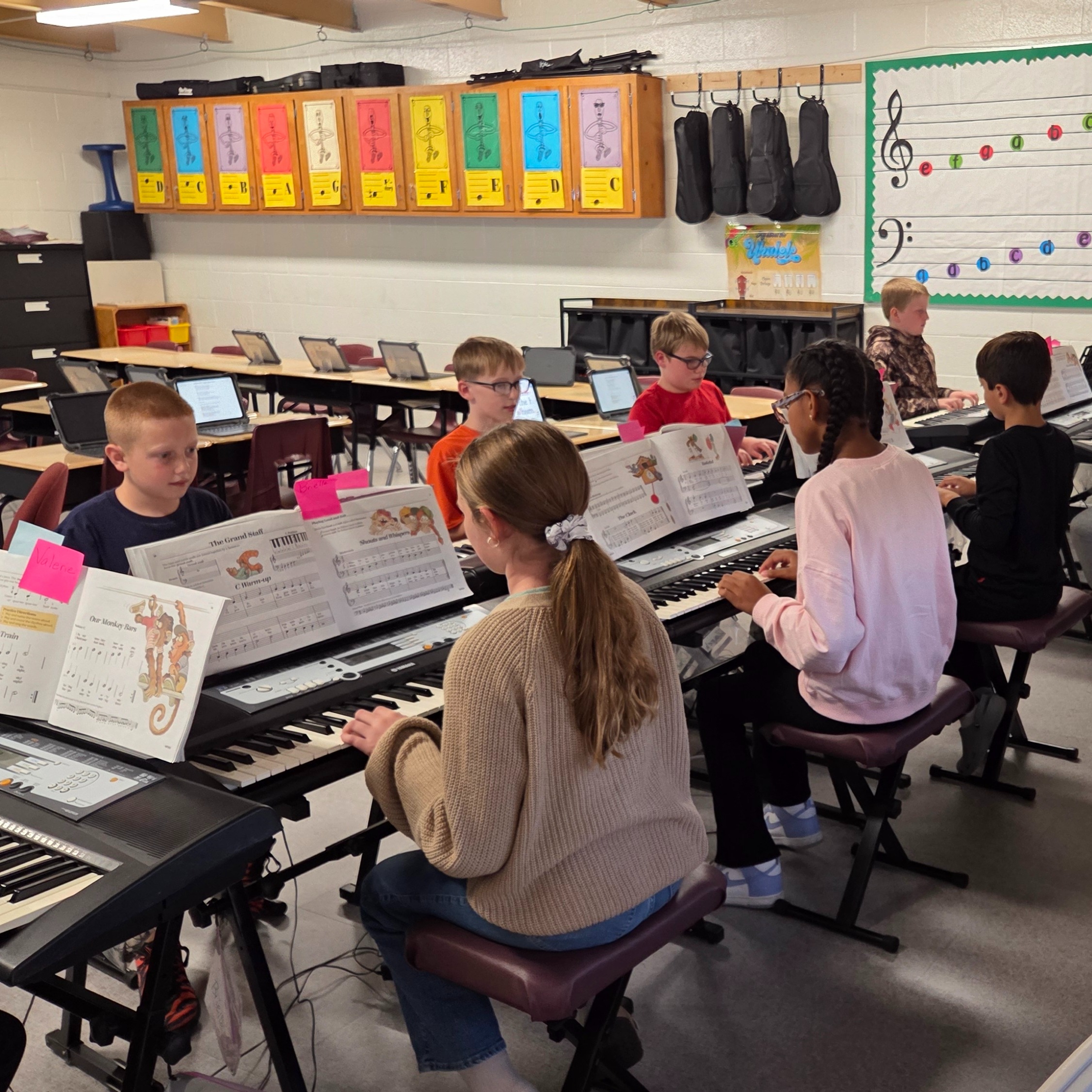 Music classroom with children sitting at keyboards. A few adults and children play music together.
