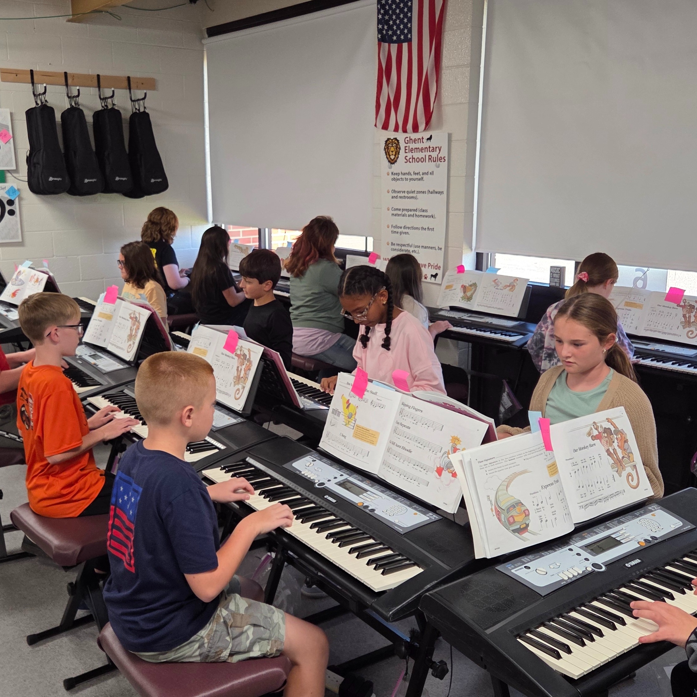 Elementary students in a classroom play keyboards and follow music books. A flag and musical notes are on the wall.