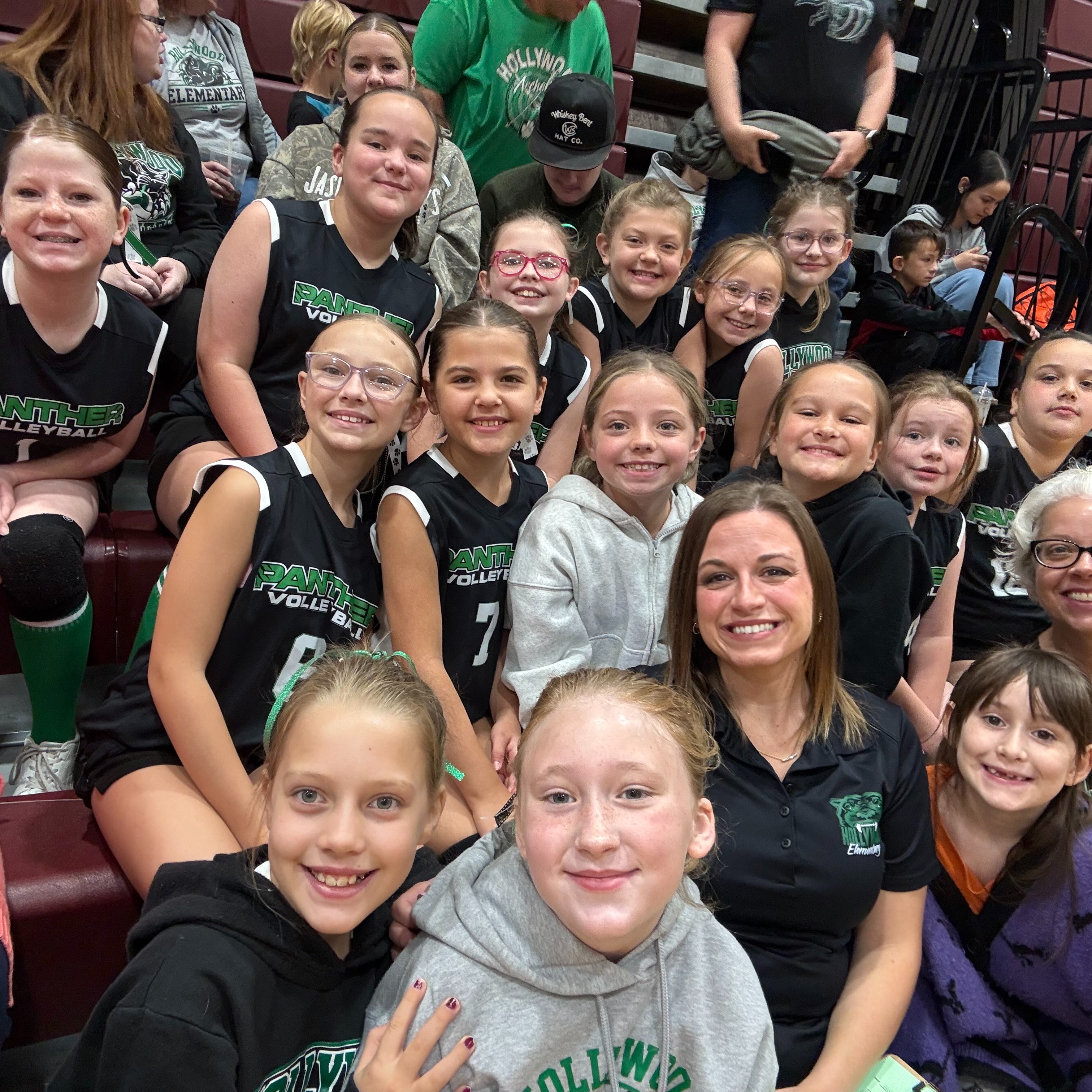 A group of young girls in sports uniforms sit on bleachers, smiling and posing for a picture.