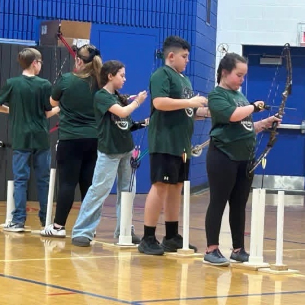 Five students stand in a gym, holding bows and arrows, with white posts in front of them.
