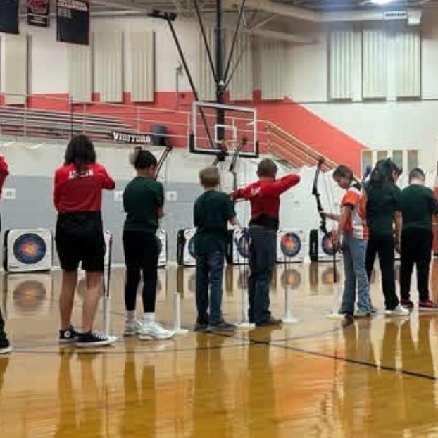 Indoor basketball court with students practicing archery. People hold bows and arrows and stand in rows.