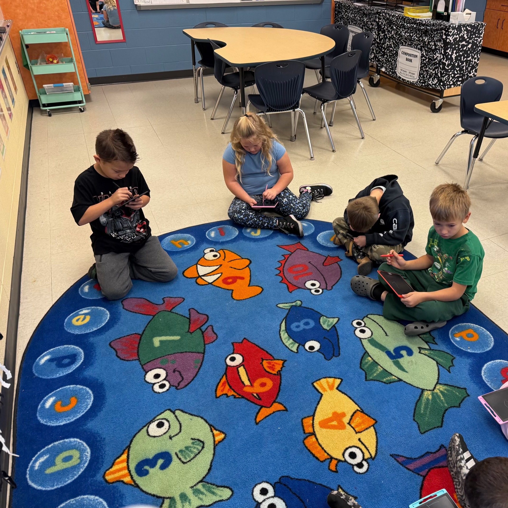 A group of young children sitting around a carpet with pictures of fish, each child using a tablet.