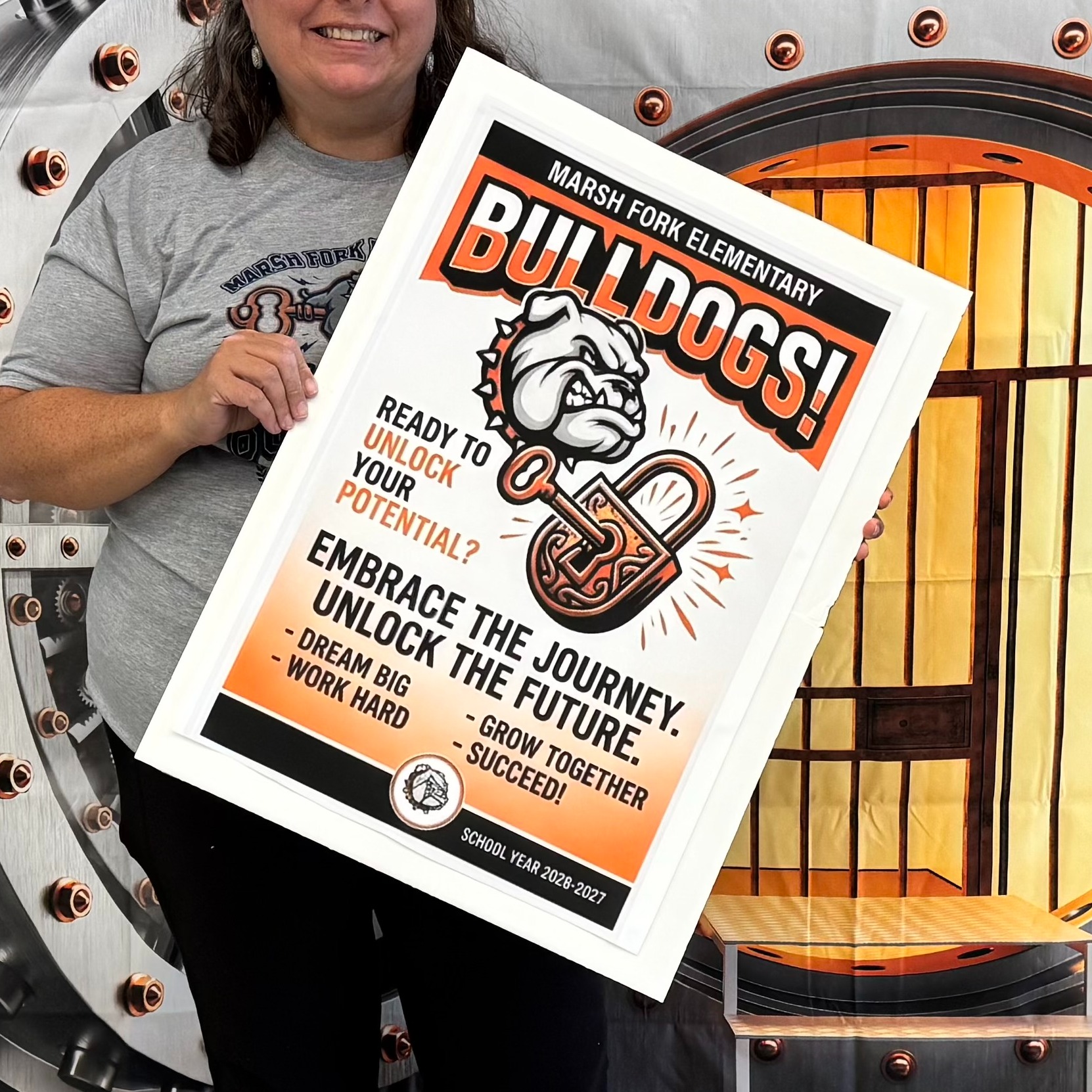A woman in a gray shirt holds a poster in front of a mock bank vault.