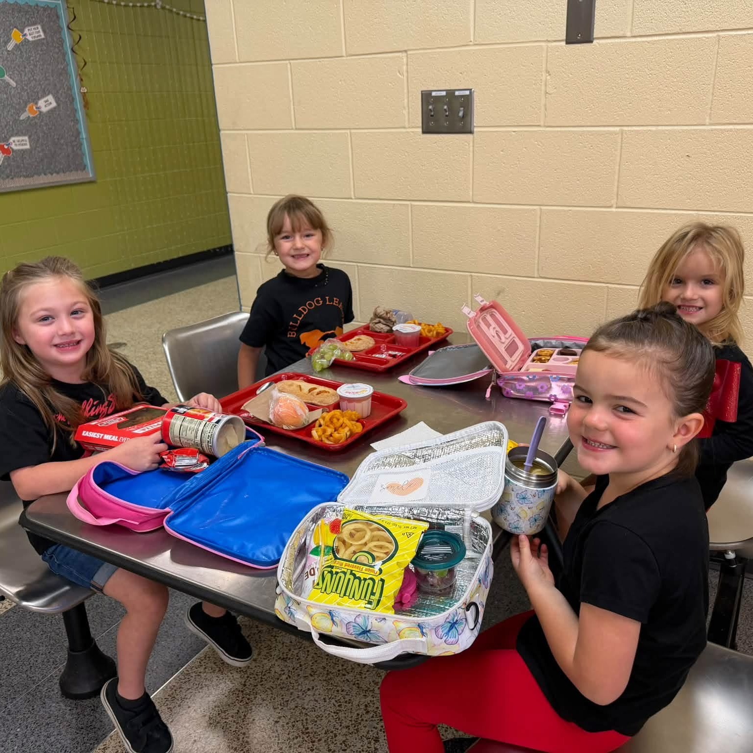 Four girls sit at a table with trays of food, holding cups and bags. A green board is behind them.