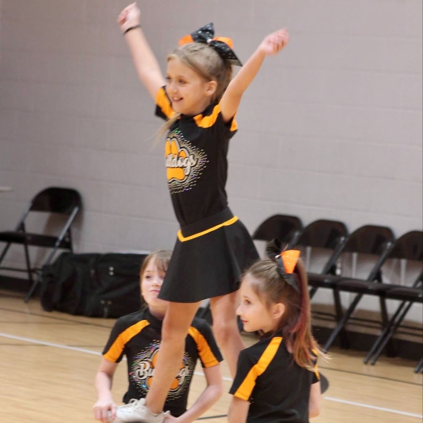 Three young girls in black and yellow cheerleading uniforms on a basketball court; one girl stands on shoulders of two others.