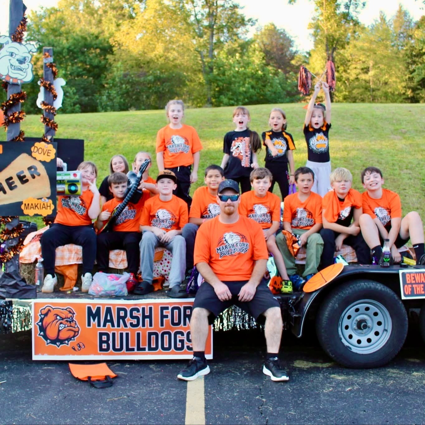 Students wearing orange shirts pose on a decorated float with orange balloons. A man sits in front.