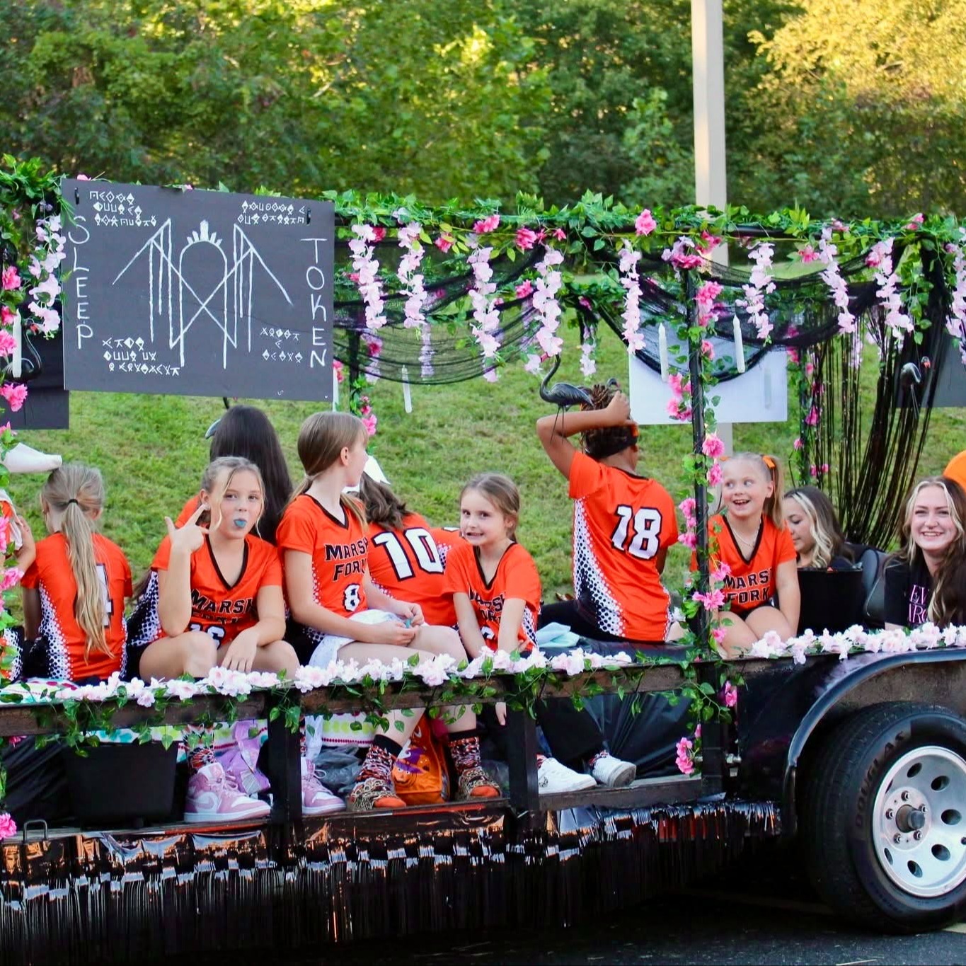 Kids in orange shirts sit on a decorated float with flowers and green leaves.