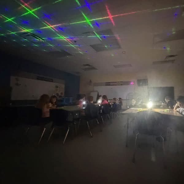Students sit around tables in a classroom. The room has colored lights on the ceiling.
