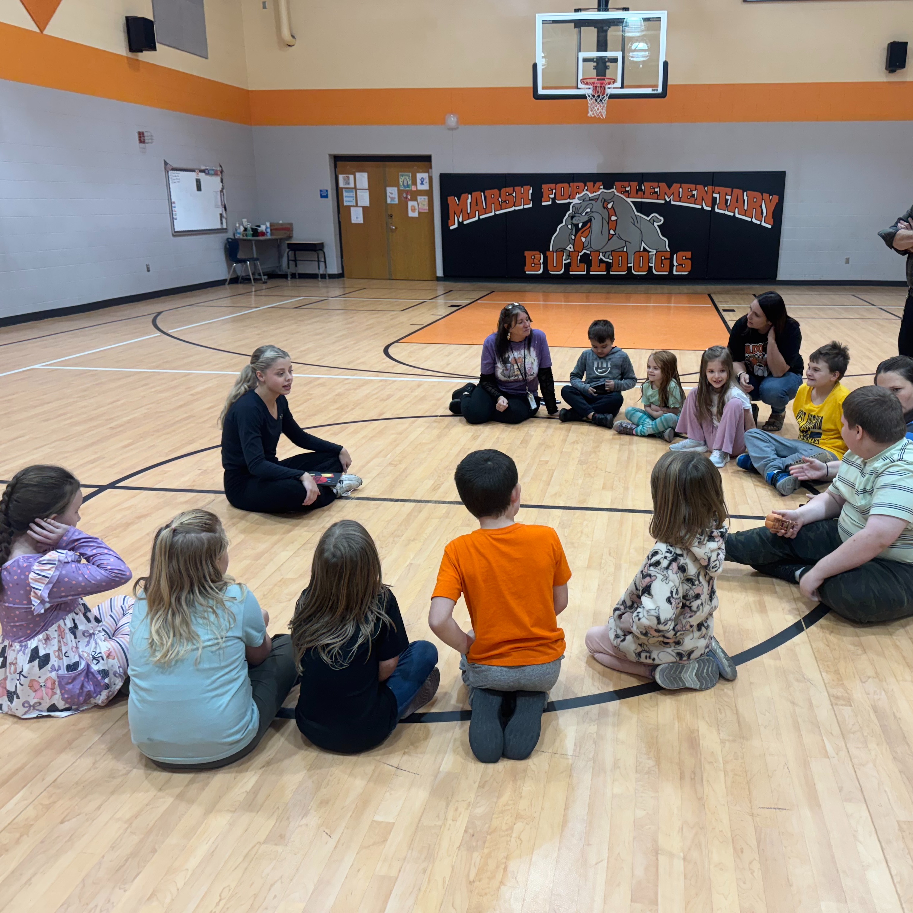 A group of children and adults sit in a circle on a basketball court. One adult stands with a clipboard.