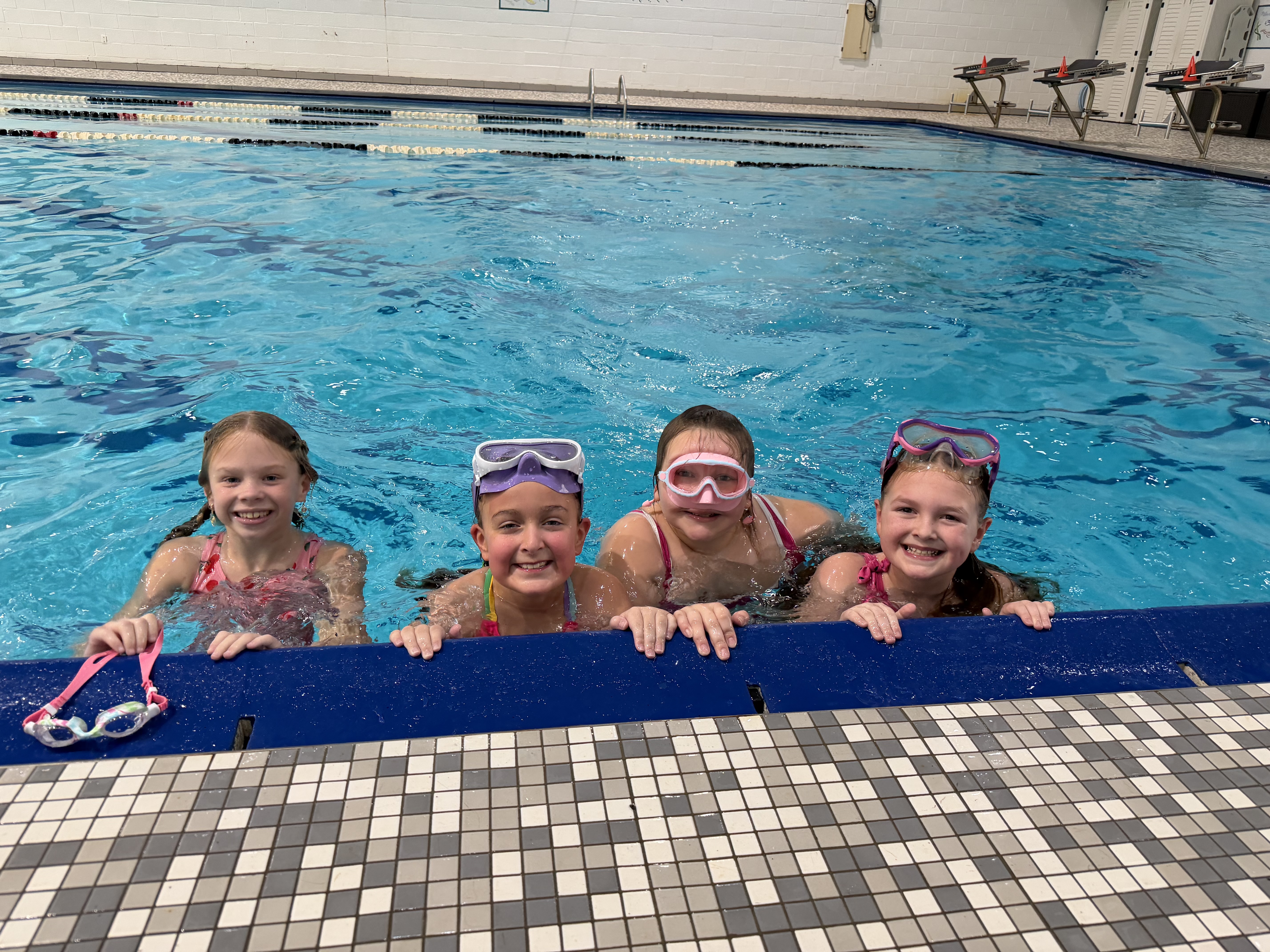 Four young girls wearing goggles sit in a pool. One holds a pair of goggles. The pool has a checkered bottom.