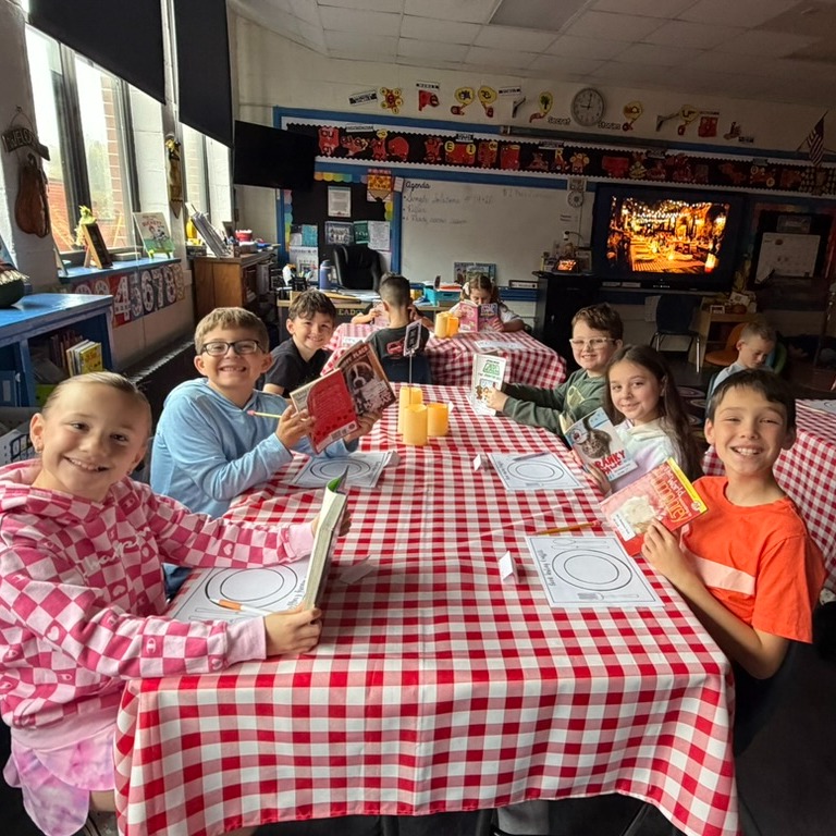 Group of students sitting at a table with red and white checkered tablecloths, holding books, in a classroom.