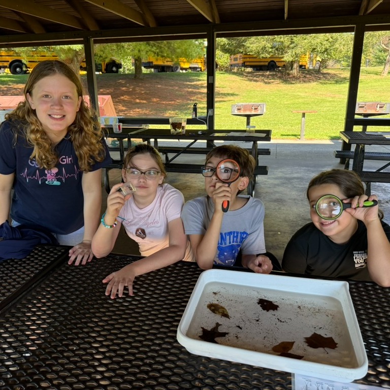 Four children wearing glasses sit at a table. A woman stands behind them. A tray is on the table.