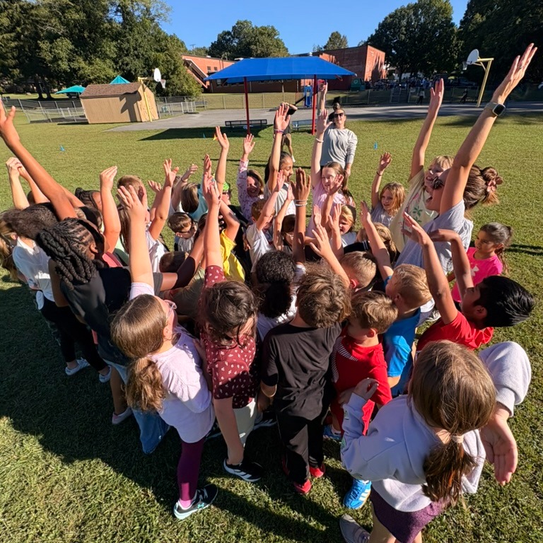 Students standing in a circle with raised hands, facing a person leading a game on a grassy field.