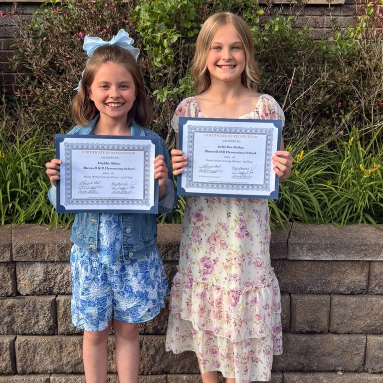 Two girls hold certificates and smile at camera; one wears sandals, the other a bow.