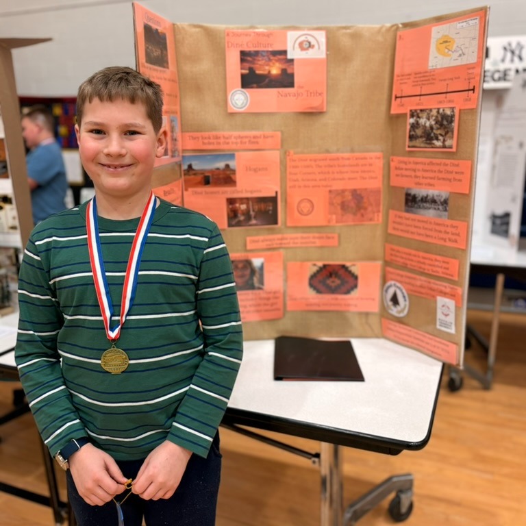 A smiling young boy with a medal stands in front of a display board with pictures.