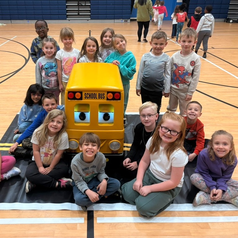 Students in a gym posing with a large yellow toy bus on a mat.