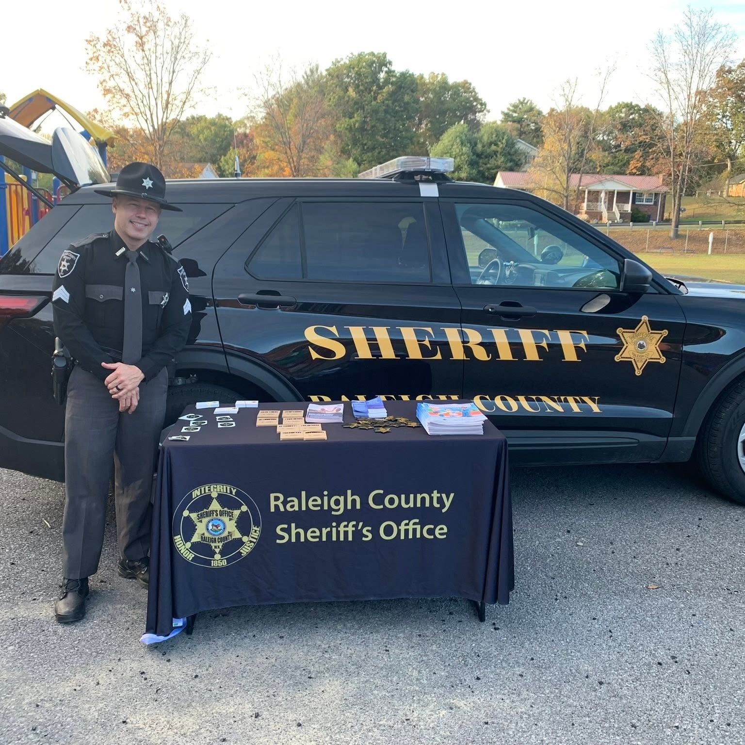 A police officer stands by a table with items. Behind him, a black SUV with "SHERIFF" and a star on the door.