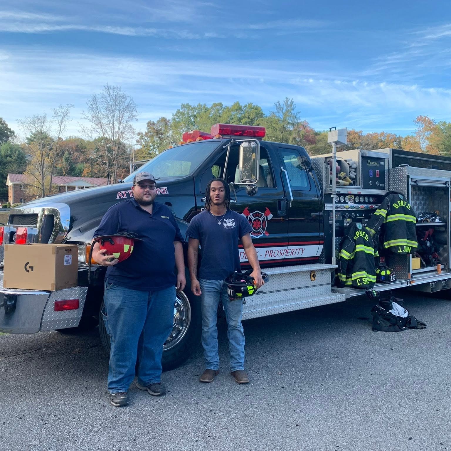 Two men stand by a firetruck with a large box in the foreground and trees in the background.