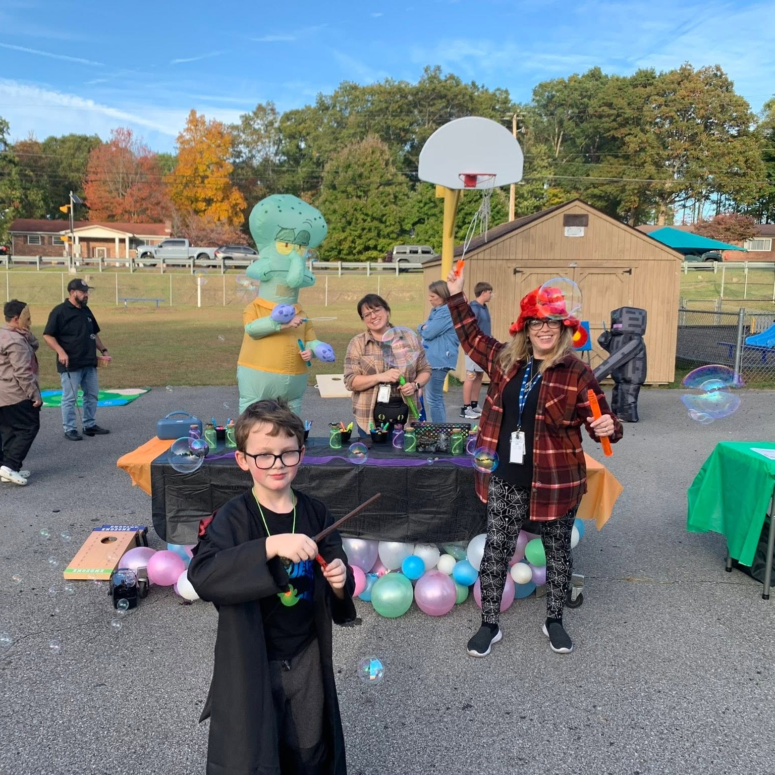 Children, adults, and mascots in a school yard. A table has balloons and a basketball hoop.