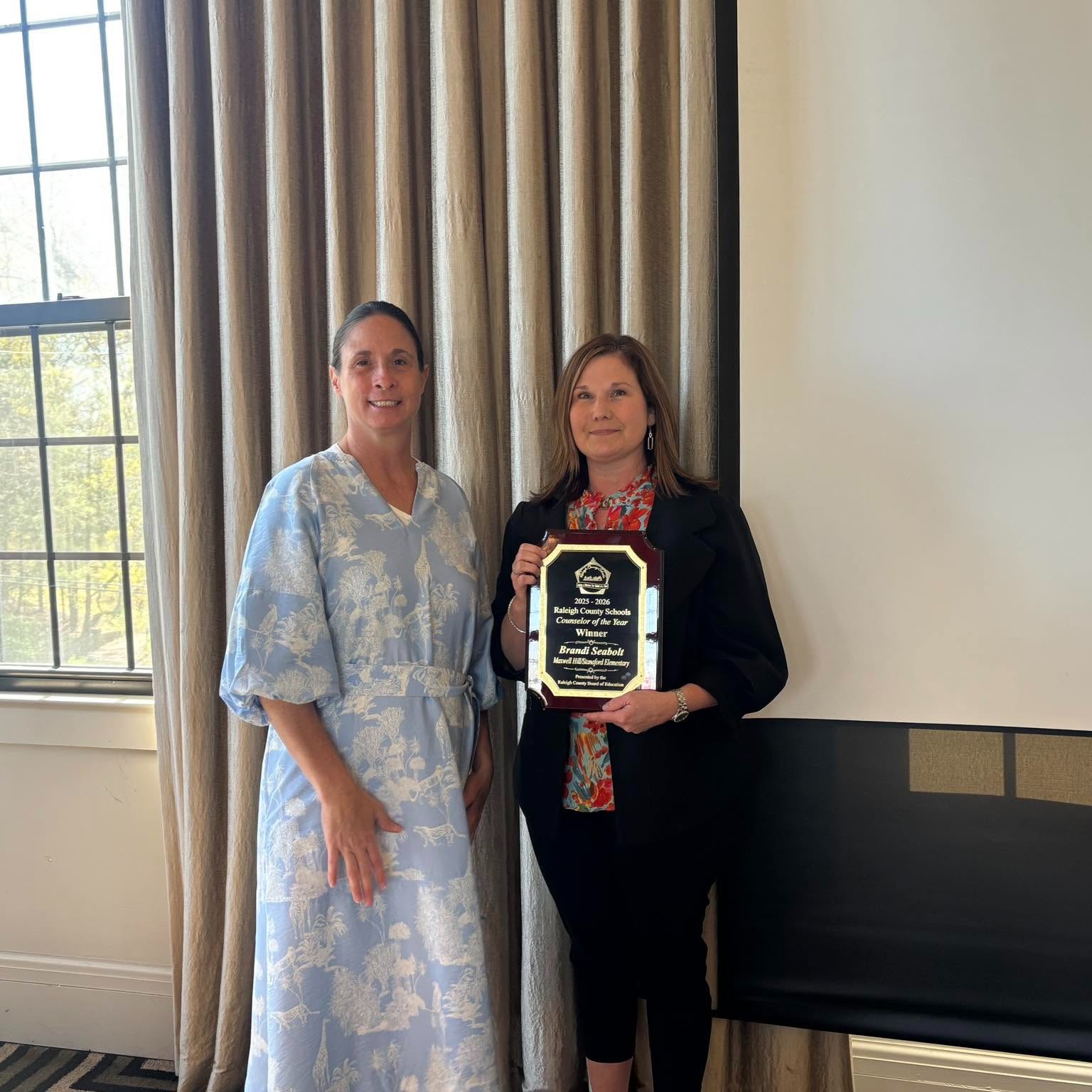 Two women in a room with a window and curtains, one holding a plaque with text and logo.