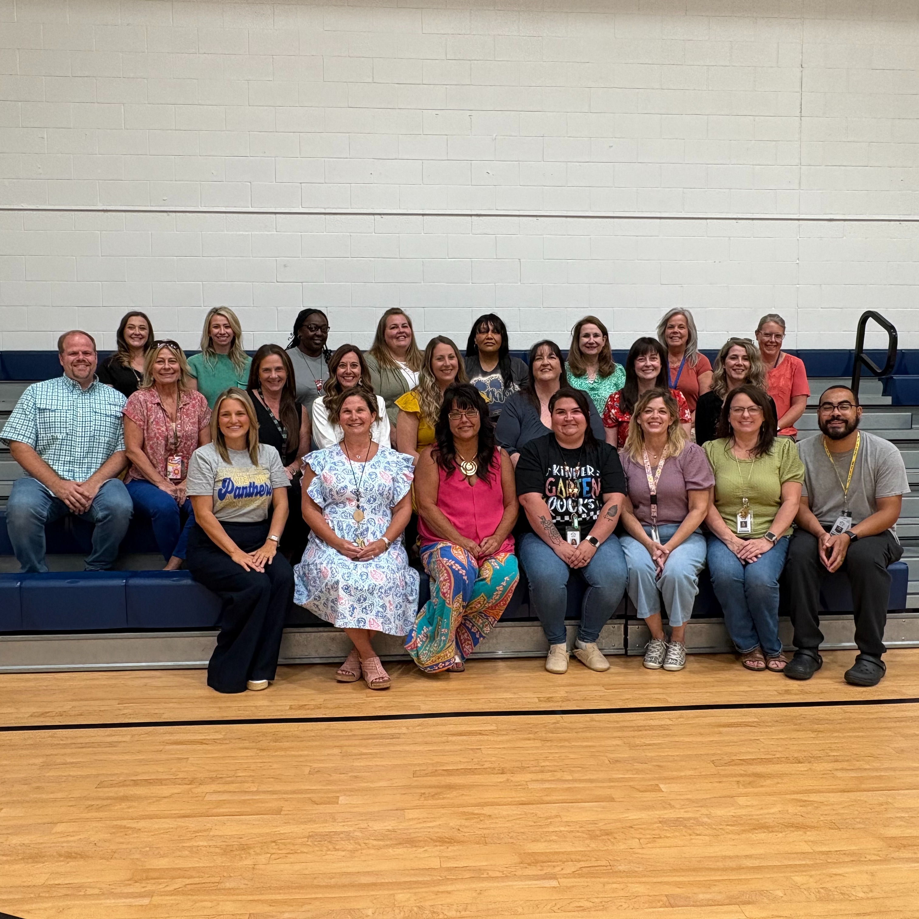 Group of adults and children sitting on bleachers in a gym with a wooden floor.
